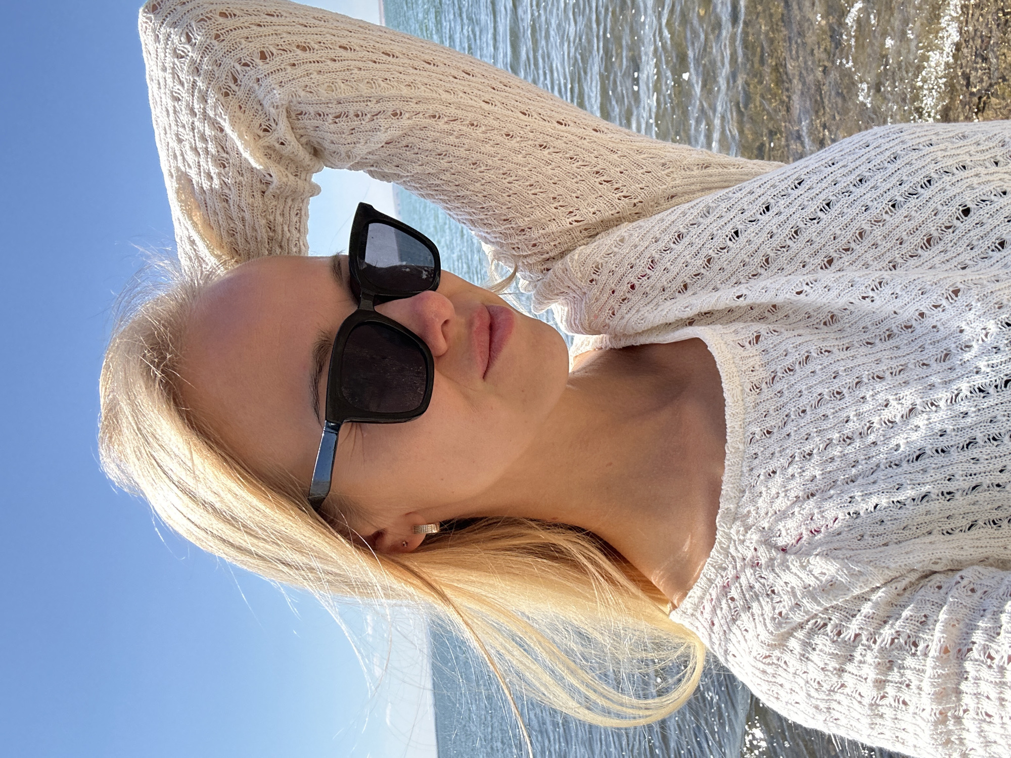 A medium shot photograph taken from a slightly low angle captures a blonde woman with fair skin standing outdoors near a body of water under a clear blue sky. The woman, positioned to the left of the frame, is wearing large, dark-rimmed sunglasses and a white, open-knit, long-sleeved sweater. Her right arm is raised, with her hand resting on the back of her head, while her left arm is lowered. Her blonde hair is styled loosely, with strands blowing in the wind. The background consists of the calm, blue water of a lake or sea, with subtle ripples and reflections of sunlight on its surface. The shoreline is visible in the lower right corner, with small rocks and pebbles. The overall impression is that of a bright, sunny day at a waterfront location. The image does not provide specific details that can be used to definitively identify the location as the city of Gornyi Erik, Russia.