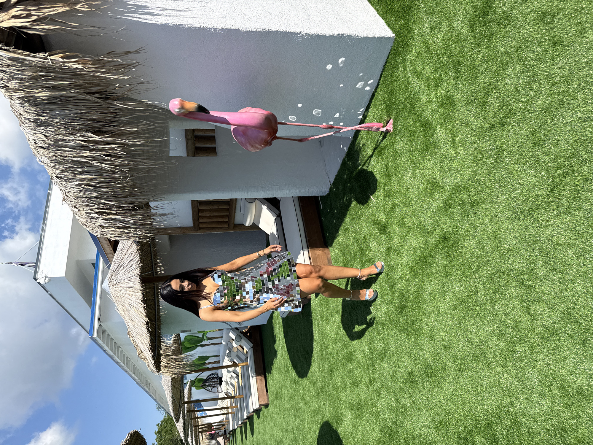 A woman walks on bright green artificial grass in front of a white building with thatched roofs in Baliovtsi, Bulgaria. She wears a short, silver, sequined dress and silver high-heeled sandals. To her right stands a pink flamingo statue. In the background, lounge chairs are visible under more thatched roofs, and trees can be seen in the distance under a clear blue sky with scattered clouds.