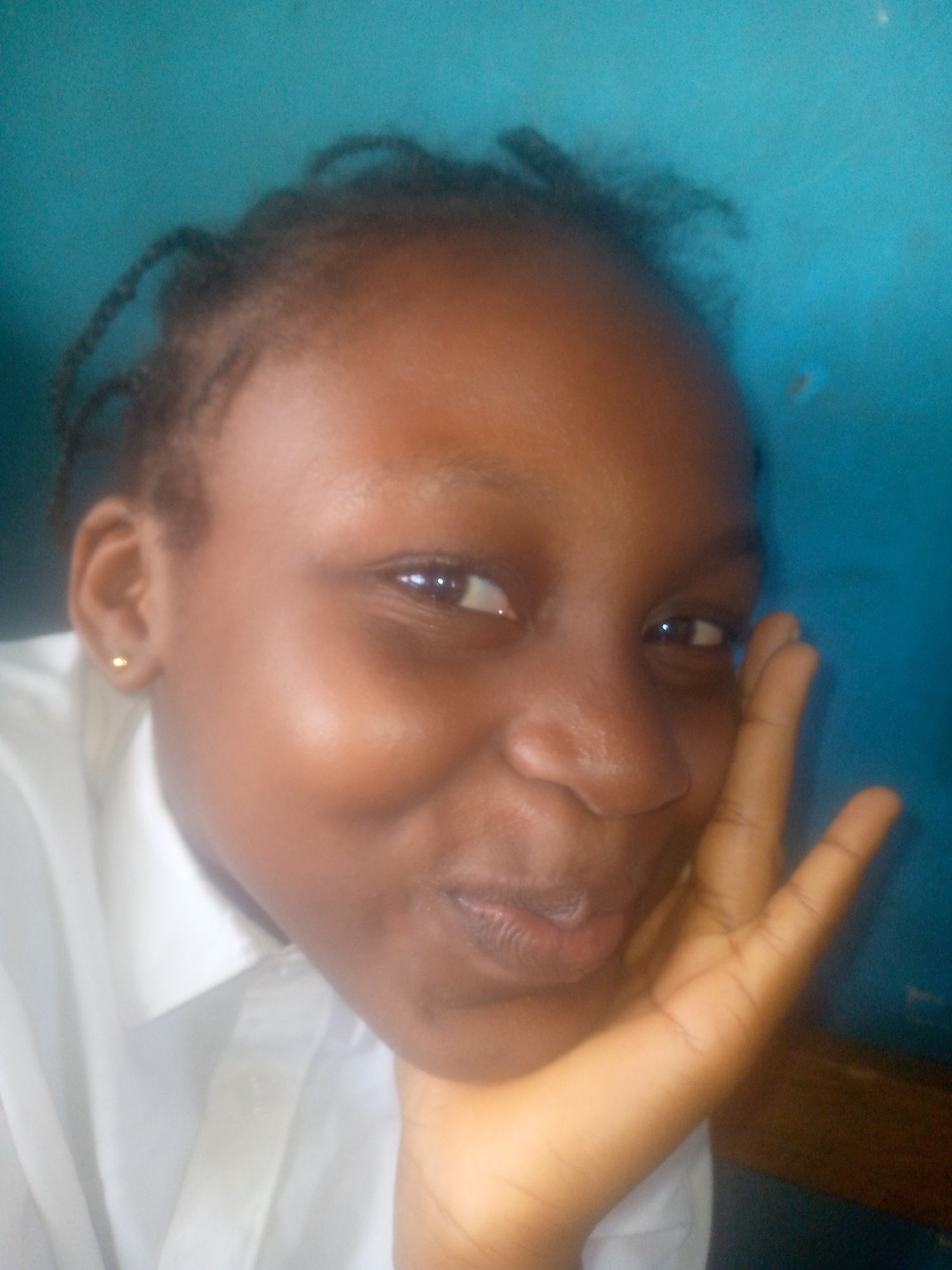 A young Black woman is posing for a selfie. She is wearing a white shirt and a gold stud earring in her left ear. Her hair is styled in small braids. She is holding her right hand up to her cheek, and she is smiling slightly. The background is a blue wall. The location appears to be indoors. There is no definitive information in the image to verify it is taken in Ikorodu, Nigeria.
