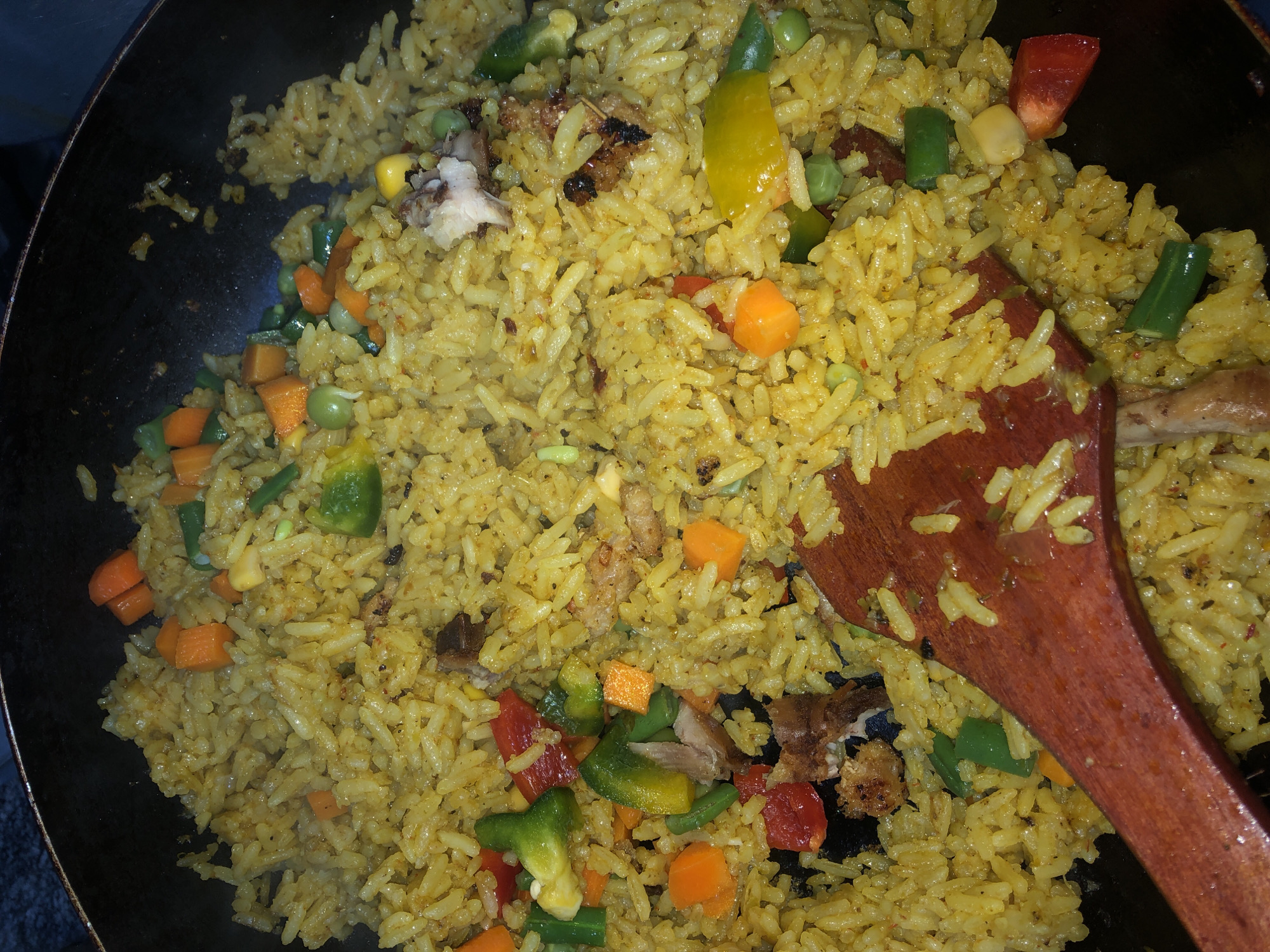 A close-up, overhead shot captures a pan filled with yellow fried rice, heavily seasoned with spices. Mixed into the rice are diced carrots, green beans, peas, corn kernels, diced red and green bell peppers, and pieces of shredded or cubed meat, some appearing slightly charred. A wooden spoon rests partially on the rice, angled from the bottom left corner of the frame. The pan itself is dark and appears to be made of cast iron or a similar material. The image is taken from a high angle, looking directly down into the pan. There is no visible information or context in the image that would allow for the identification of a specific location within Bar Jirgi Summa, Nigeria, or any other geographical area.