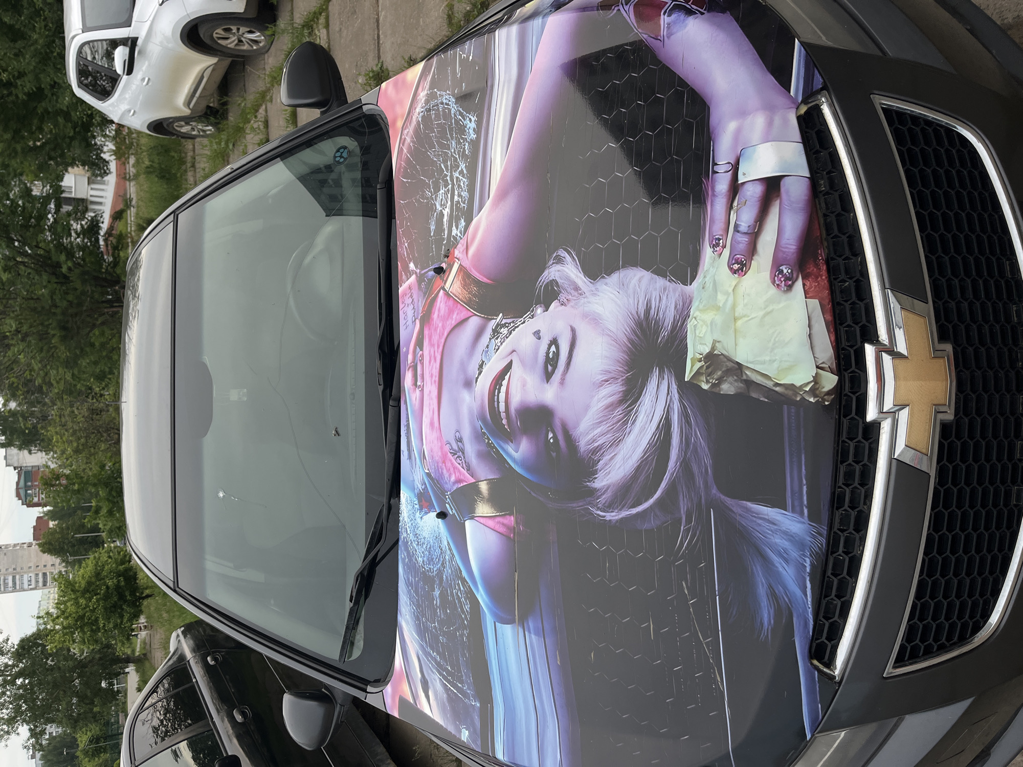 An outdoor, eye-level, side-angle shot captures a dark-colored Chevrolet car with a vibrant custom hood wrap, parked in what appears to be an urban or suburban setting in Chaikovsky, Russia. The image is oriented vertically, showing the car's front and left side.
The dominant feature is the car's hood, adorned with a large, colorful graphic of a female character strongly resembling Harley Quinn. She is depicted with light-colored hair, a wide, joyful smile, and is looking directly towards the viewer. Her left arm and hand are visible, featuring several rings, a wide silver bracelet or cuff, and glitter-decorated fingernails. She holds a crumpled yellow cloth or paper in her hand. The background of the wrap displays a dark, honeycomb-like geometric pattern interspersed with splashes of pink, blue, and purple. Notably, there is a visible section of cracking or peeling on the wrap near the windshield on the left side, resembling shattered glass.
The car's windshield shows minor marks or debris. A portion of the Chevrolet emblem is visible on the grille. In the background, a white SUV is partially visible parked behind the main car, with green trees and distant residential buildings under what appears to be a daytime sky. No specific activity is occurring, the car is stationary.