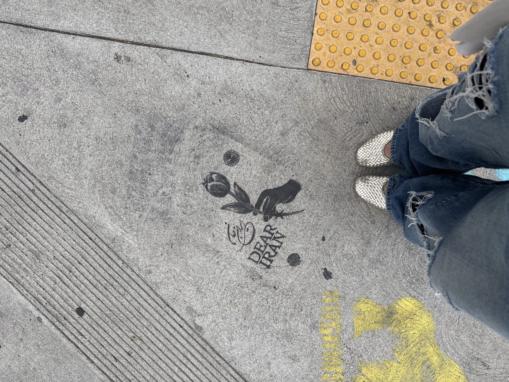 The image shows a concrete sidewalk in Los Angeles, California. The sidewalk is textured and has a few visual elements: tactile paving with raised circular dots in yellow, several sets of horizontal lines in the concrete, and a dark-colored stencil with a tulip, a hand holding the tulip, and the text "DEAR IRAN." There are also some small dark splotches on the concrete. A person is standing on the sidewalk. The person is wearing blue jeans with large tears and silver woven shoes. Yellow text is visible on the sidewalk edge and is partially obscured.