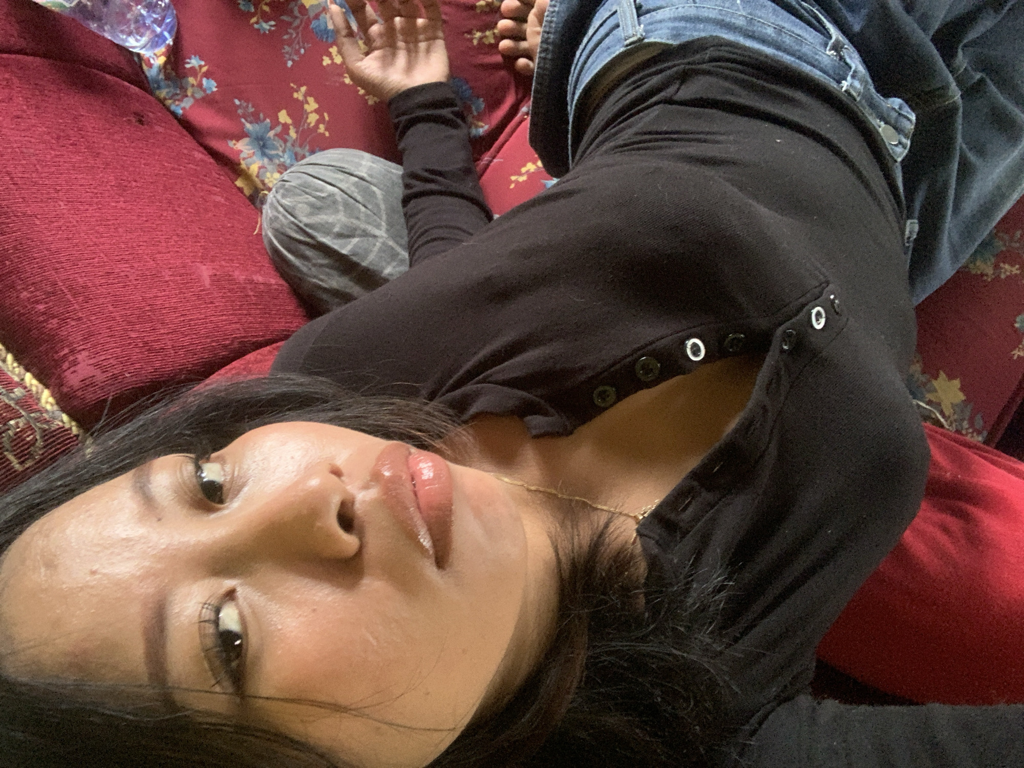 A young Asian woman with dark hair and brown eyes is lying on a red couch in Jajag, Indonesia. She is wearing a black long-sleeved shirt with buttons down the front and blue jeans. Her face is close to the camera, and she is looking off to the side. Her lips are glossy, and she has on a light amount of makeup. The couch is made of a textured fabric and has a floral pattern on the armrest. There is a bottle of water on the couch next to her. The lighting in the image is soft, suggesting it was taken indoors.
