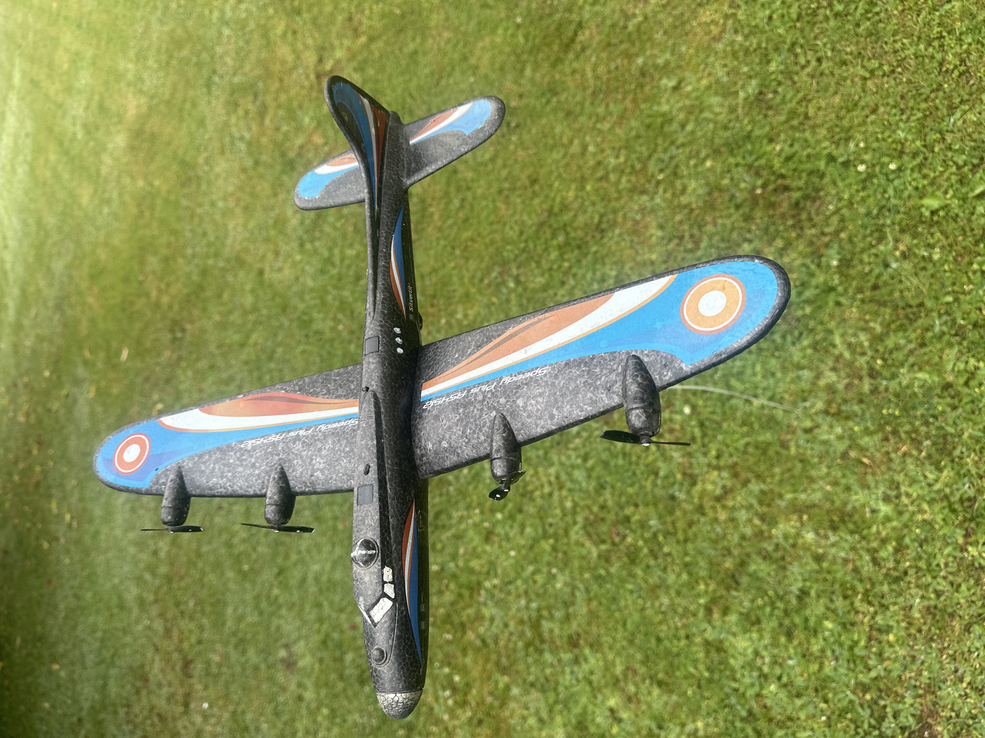 A black toy airplane is positioned outdoors on a patch of green grass. The airplane has four propellers, two on each wing. The wings and tail are decorated with blue and orange stripe patterns and circular insignia. The scene appears to be in Lapės, Lithuania, as indicated by the provided metadata. The grass is well-maintained and appears to be a lawn. The lighting suggests it is daytime, with no specific weather conditions discernible. There are no people or animals visible in the image.