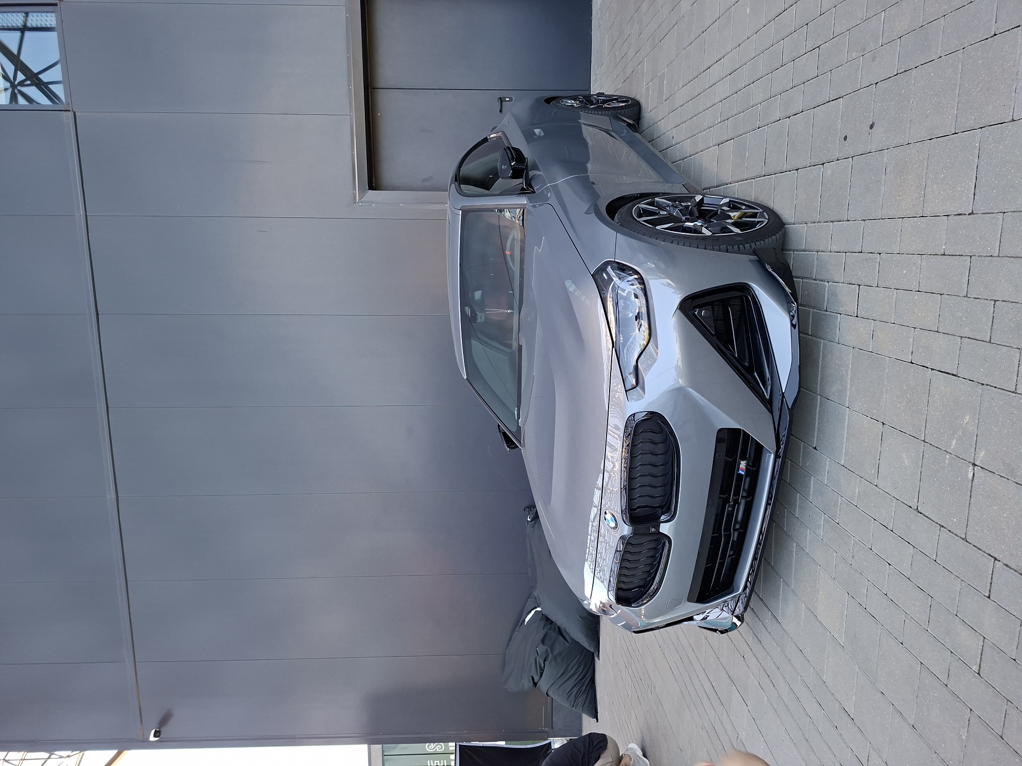 A silver BMW M2 Coupe is parked on a paved area in front of a large, dark gray modern building. The building has a large glass section at the top left and a pair of large gray doors on the right. The car is angled towards the viewer, with its front end slightly to the left. The car has a sporty body kit, including a prominent front splitter and large wheels. Behind the car, two large, black beanbag chairs are placed against the building wall. In the lower left of the frame, a person's legs and feet, wearing light-colored pants and white sneakers, are visible, suggesting they are crouching or kneeling. Another person's bare arm is visible in the lower left corner of the frame. The ground is paved with rectangular gray interlocking bricks. The scene appears to be outdoors, possibly at an event or a car dealership.