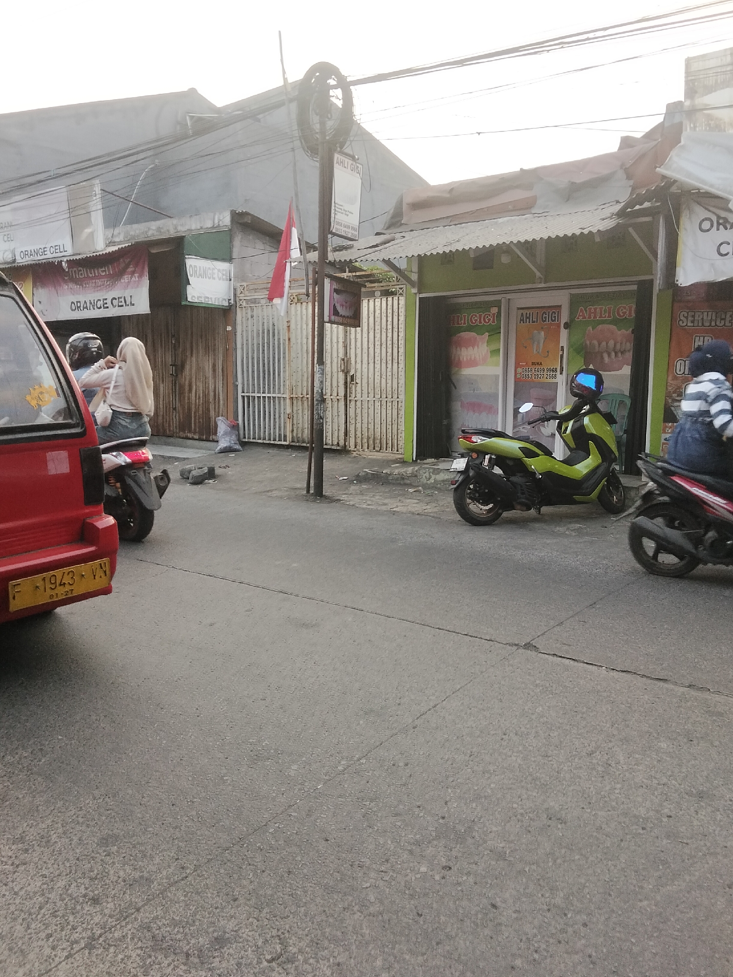 The image depicts a street scene in Cisaat, Indonesia. On the left, the rear of a red car with license plate F 1943 VN is visible. To the right of the car, two individuals on a scooter are seen, with one wearing a light-colored hijab. A green scooter is parked in front of a building with signs for "AHLI GIGI" (Dentist). To the right of the green scooter, another scooter with a person wearing a striped shirt is partially visible. The buildings in the background have corrugated metal roofs and some signage, including "ORANGE CELL". The environment appears to be a commercial street with various shops.