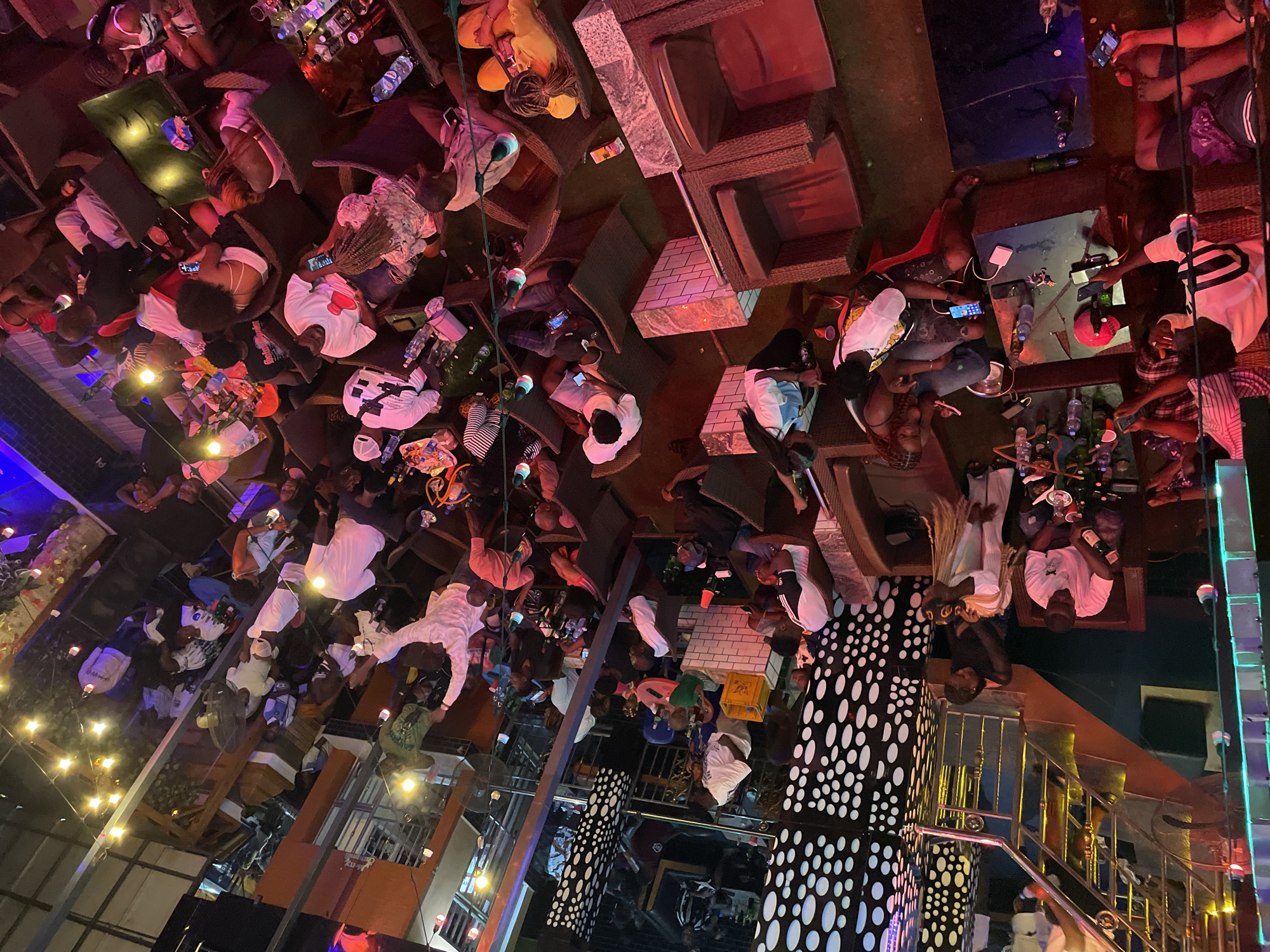 The image depicts an overhead view of a crowded indoor entertainment venue, likely a nightclub or lounge, in an unspecified city. Patrons are seated at tables and in lounge chairs, many holding cell phones. The environment is dimly lit, with artificial lighting from overhead fixtures and string lights creating a vibrant atmosphere.
In the foreground, several individuals are visible. A man in a white shirt with black stripes sits at a low table, holding a bottle. To his right, a person in a white outfit with blonde braids is also seated. Further to the right, another individual is looking at their phone. In the lower left corner, a person in a dark shirt and shorts is lying down.
The space features a mix of seating arrangements, including dark wicker lounge chairs and low tables. The decor includes a distinctive black and white speckled pillar or wall feature. A staircase with a metal railing is visible in the upper left portion of the image. Bottles of beverages and glasses are present on some of the tables, suggesting a social gathering. The overall impression is one of a lively evening event with many people engaged in socializing and using their electronic devices.
