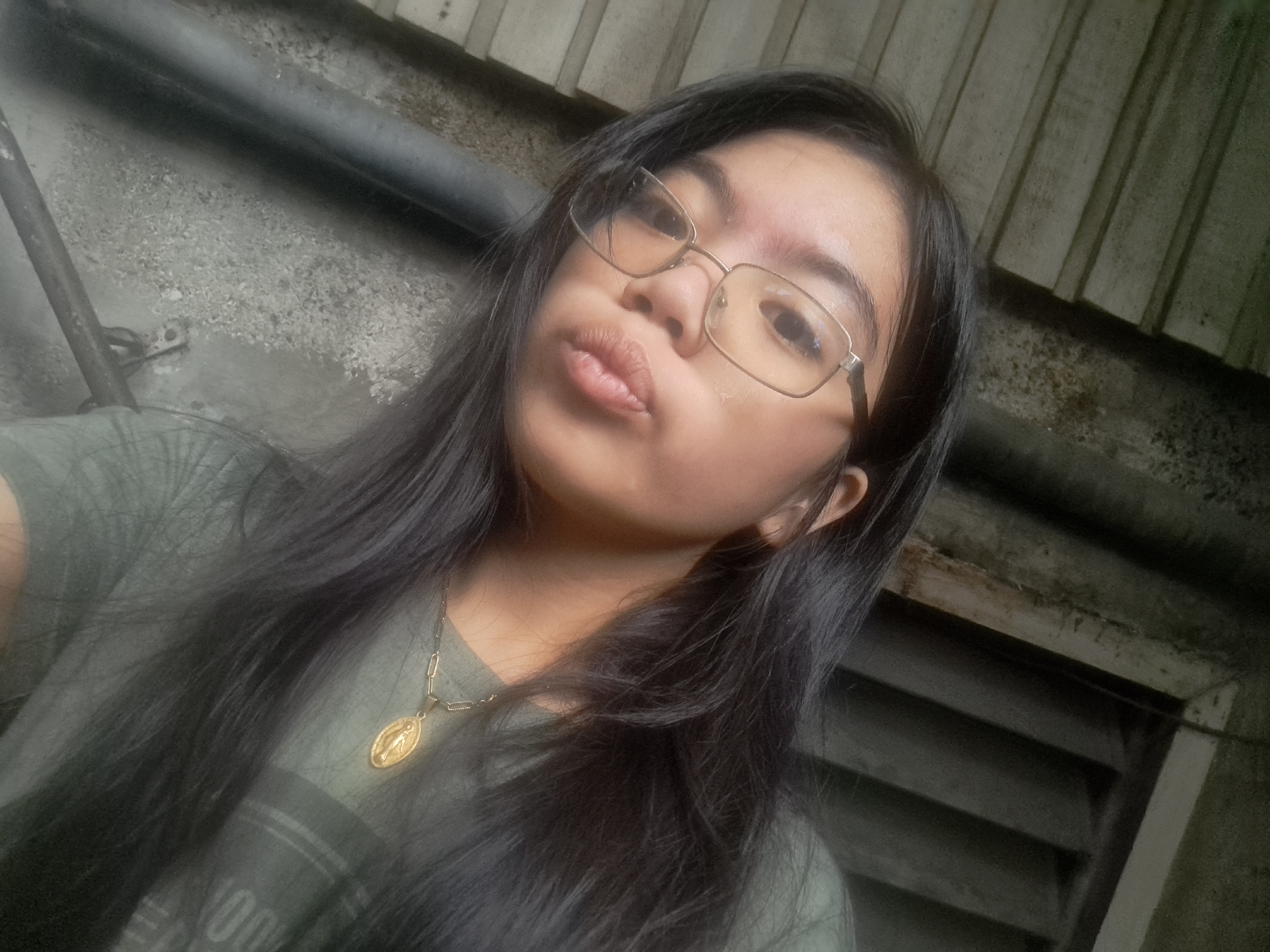 A young woman with long, dark hair and glasses is taking a selfie. She is wearing a green t-shirt and a gold necklace with a pendant. The background appears to be an outdoor setting with a concrete wall and a corrugated metal roof. The lighting suggests it is daytime, possibly overcast. The woman is in Mandaluyong City, Philippines.
