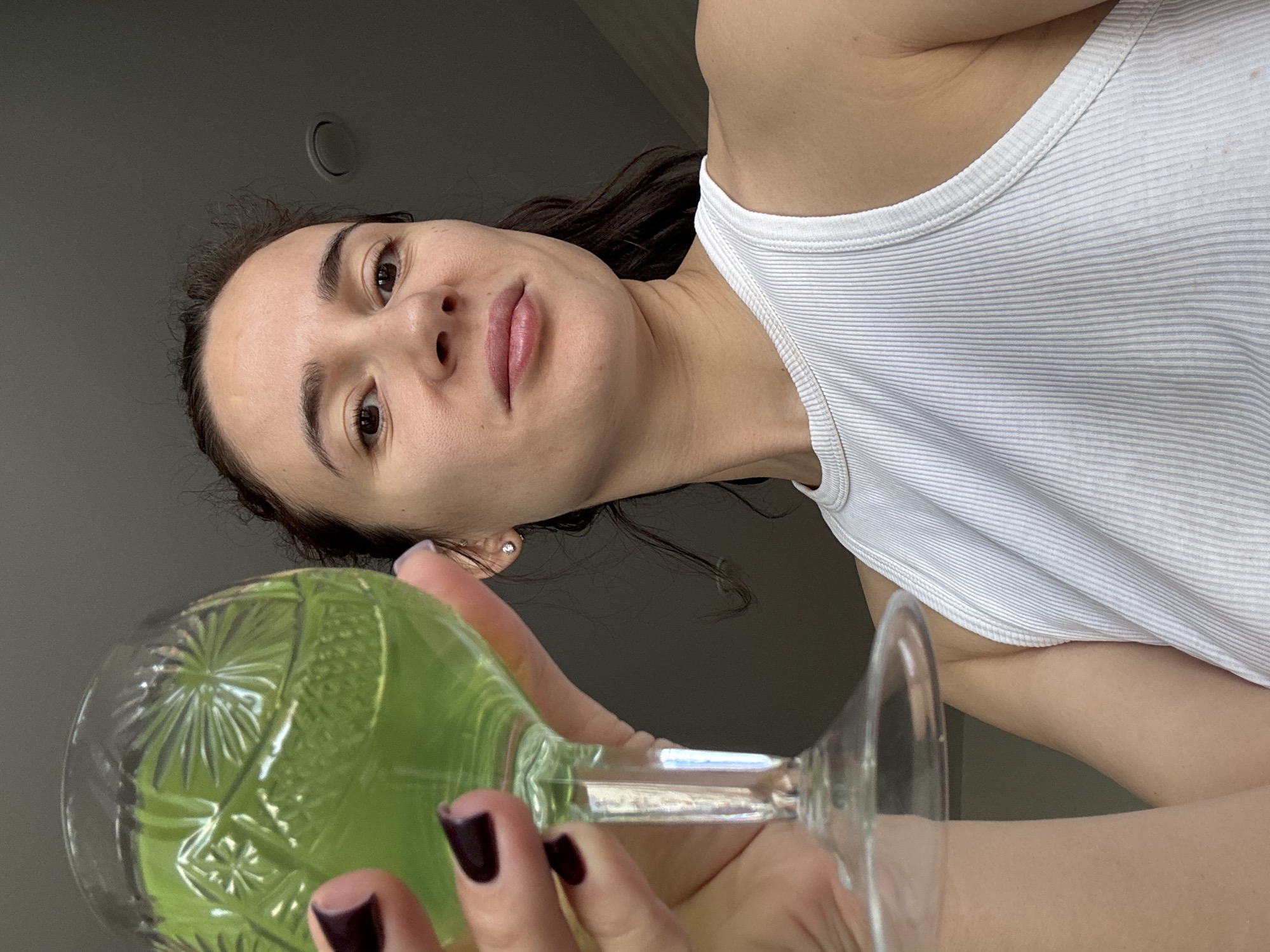 A person with light skin, brown hair, and dark burgundy nail polish holds a patterned glass filled with a green liquid. The person is wearing a white ribbed tank top. The background is a plain, light gray wall. A ceiling light fixture is visible in the upper left corner. There is no evidence suggesting the location is Chaikovsky, Russia.