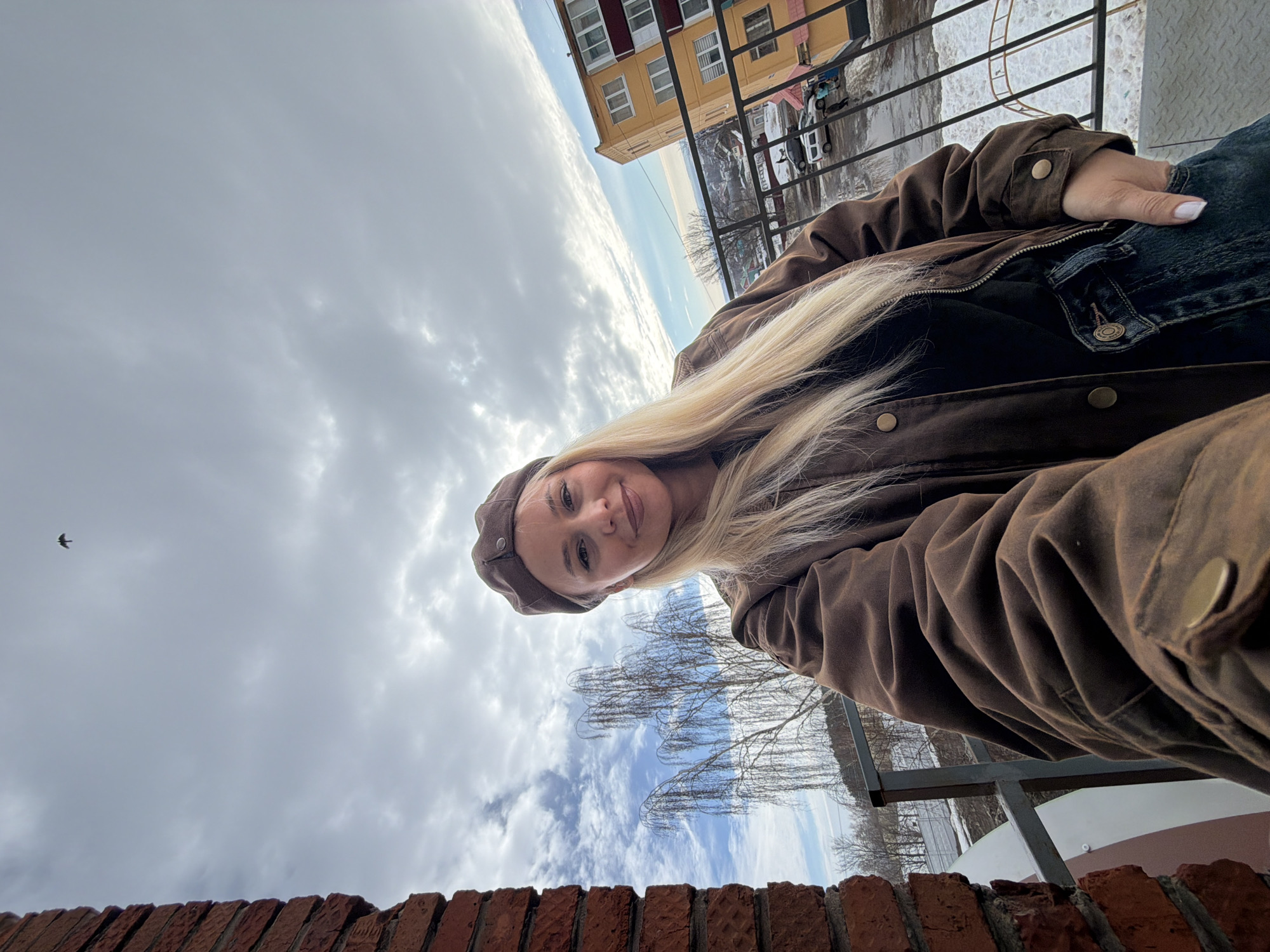 A blonde-haired woman, wearing a brown beret, a brown jacket, and blue jeans with her right hand in a pocket, smiles slightly while taking a selfie from a balcony in Sarapul, Russia. A red brick wall is visible on the left. Beyond a black metal railing, the background reveals a cityscape under a partly cloudy sky with a single bird in flight near the top left. A bare, weeping-type tree is prominent in the mid-ground. Further in the distance, a multi-story yellow building with white windows and red accents is visible, along with other indistinct structures. The ground below consists of a wet street with parked vehicles and residual patches of snow. Several indistinct figures are present on the street level.