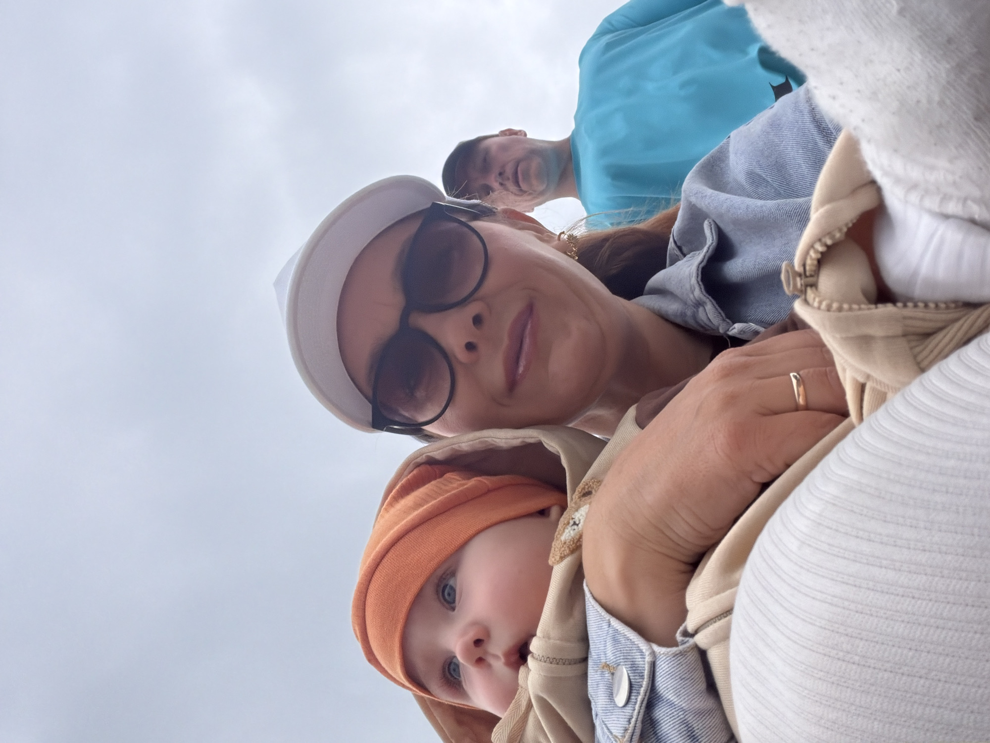 This is a vertical, eye-level, close-up, outdoor photograph taken from a low angle looking up. The sky is overcast and light gray, suggesting cloudy weather and possibly a cooler day. In the foreground, a baby with bright blue eyes is swaddled in a tan hooded garment and an orange beanie, peeking out from behind an adult. The baby's face is the primary focus, with its gaze directed slightly off-camera. To the left of the baby, a woman is visible, wearing dark sunglasses, a white cap, and a denim jacket. Her hand, adorned with a gold ring, rests on the baby's garment. Behind the woman, a man wearing a bright blue shirt is partially visible, looking towards the camera. The background is filled with the pale sky. The overall impression is of a casual, family outing, possibly during the day, given the lighting. The location context provided is Chaikovsky, Russia. There is no visible text in the image.