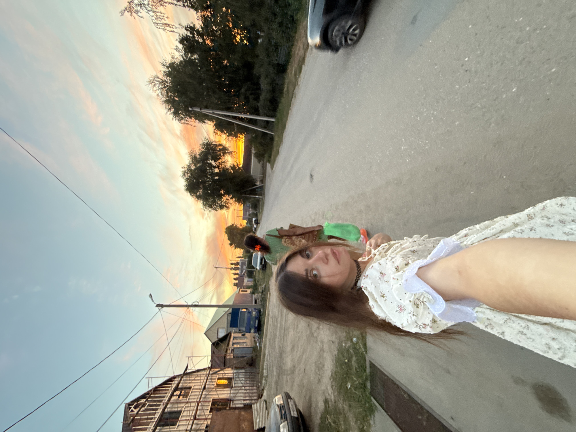 An outdoor, low-angle, medium shot captures a young woman in the foreground, wearing a white floral dress and a black choker, turning to look back at the camera with a neutral expression. Her right arm is extended forward, holding a green plastic bag. Behind her, another woman in a green dress, carrying a patterned bag and a green plastic bag, walks away from the camera down a paved road. The scene is set in a rural or semi-rural area, with houses and trees lining the road. The sky above is a vibrant mix of blues and oranges, indicating a sunset. Power lines are visible stretching across the sky. A black car is parked on the right side of the road, and other vehicles and structures are visible in the background. The setting appears to be Sol-Iletsk, Russia, given the architectural style and general atmosphere.