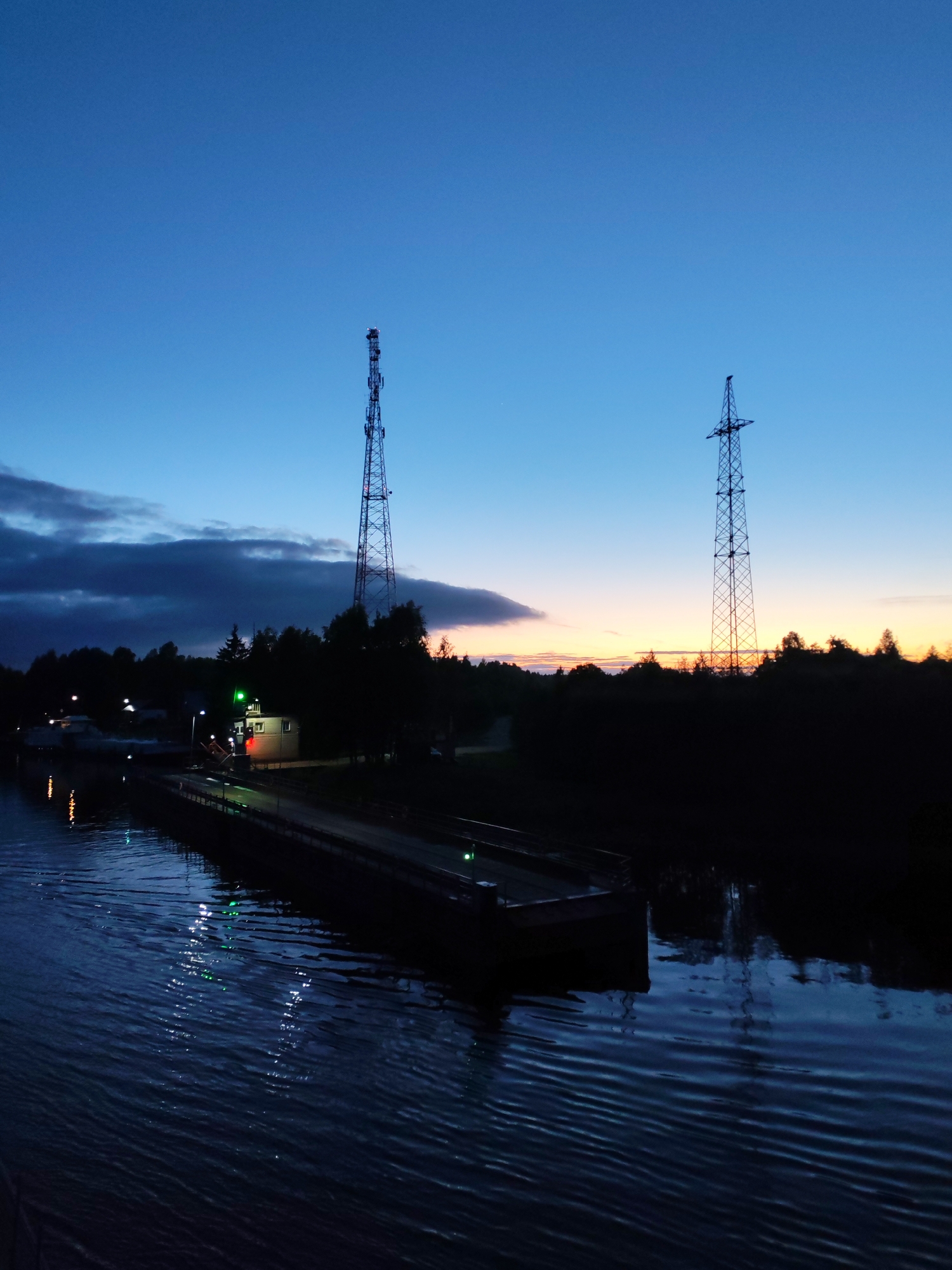 The image shows Annensky Most, Russia at twilight. The sky is a gradient of deep blue to a pale orange on the horizon. Two lattice towers stand silhouetted against the sky. A dark, elongated object, possibly a barge, is visible in the water with green lights reflected. A small building with green lights is situated near the barge. A bank of trees and dark clouds are positioned to the left. The water surface is rippled.