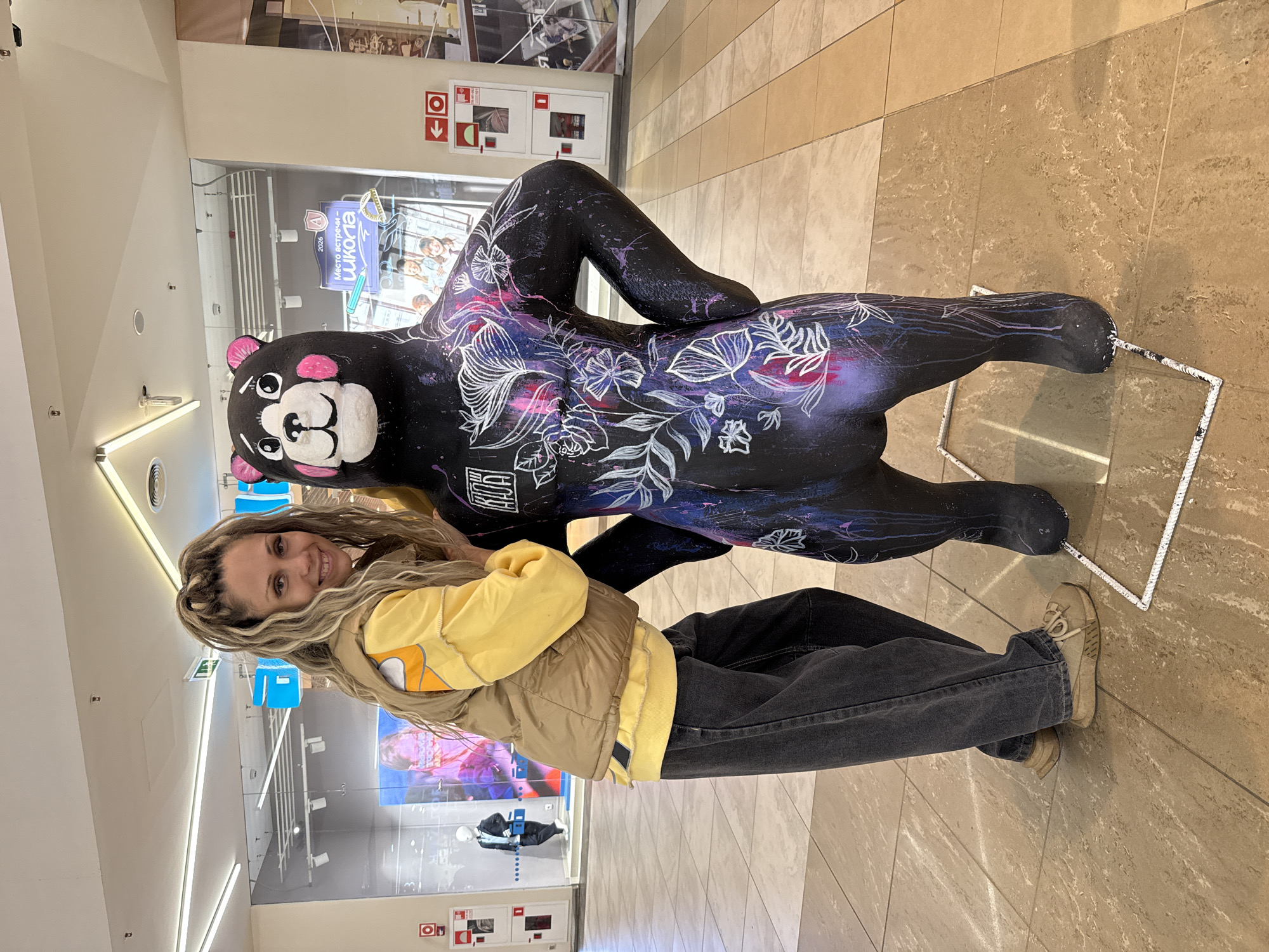 A woman with blonde dreadlocks stands next to a life-sized statue of a black bear decorated with white and pink floral patterns. The woman is wearing a yellow long-sleeved shirt, a beige vest, and dark gray wide-leg jeans. She is smiling at the camera and has her left arm resting on the bear's body. The bear statue is standing upright with its hands on its hips. In the background, there are store windows and mall lighting. One window displays a mannequin and a poster with Russian text that reads "Место встречи – школа" (Meeting place - school). The floor is tiled in a light brown and beige pattern. The scene appears to be in a shopping mall in Yaroslavl, Russia.