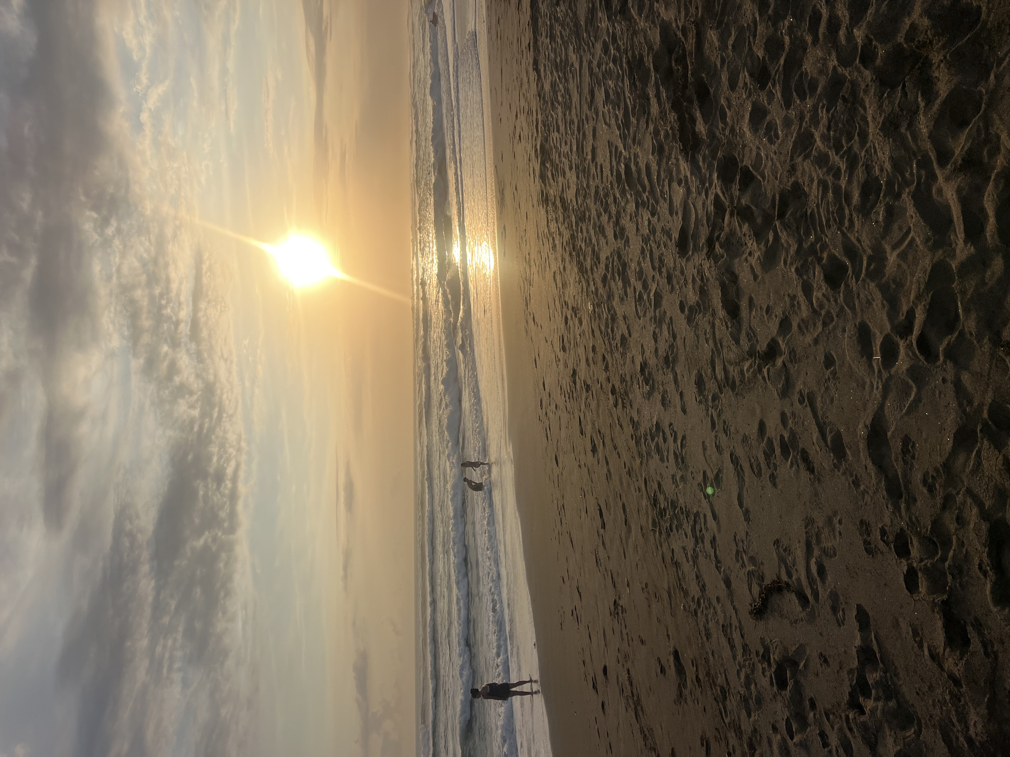 The image depicts a sunset scene at a beach, with the sun casting a warm glow over the water and sky. The sky is filled with scattered clouds, and the sun itself is a bright orb with a prominent lens flare. Gentle waves are rolling onto the sandy shore, where several people are present. One person stands near the water's edge on the left, facing the ocean. Further out in the water, two other figures are visible, appearing to be engaged in some activity. The foreground is dominated by the textured sand of the beach, with numerous footprints indicating recent activity. The overall atmosphere is tranquil and serene. The specific location of Kediri, Indonesia, is not directly identifiable from visual cues within the image itself.