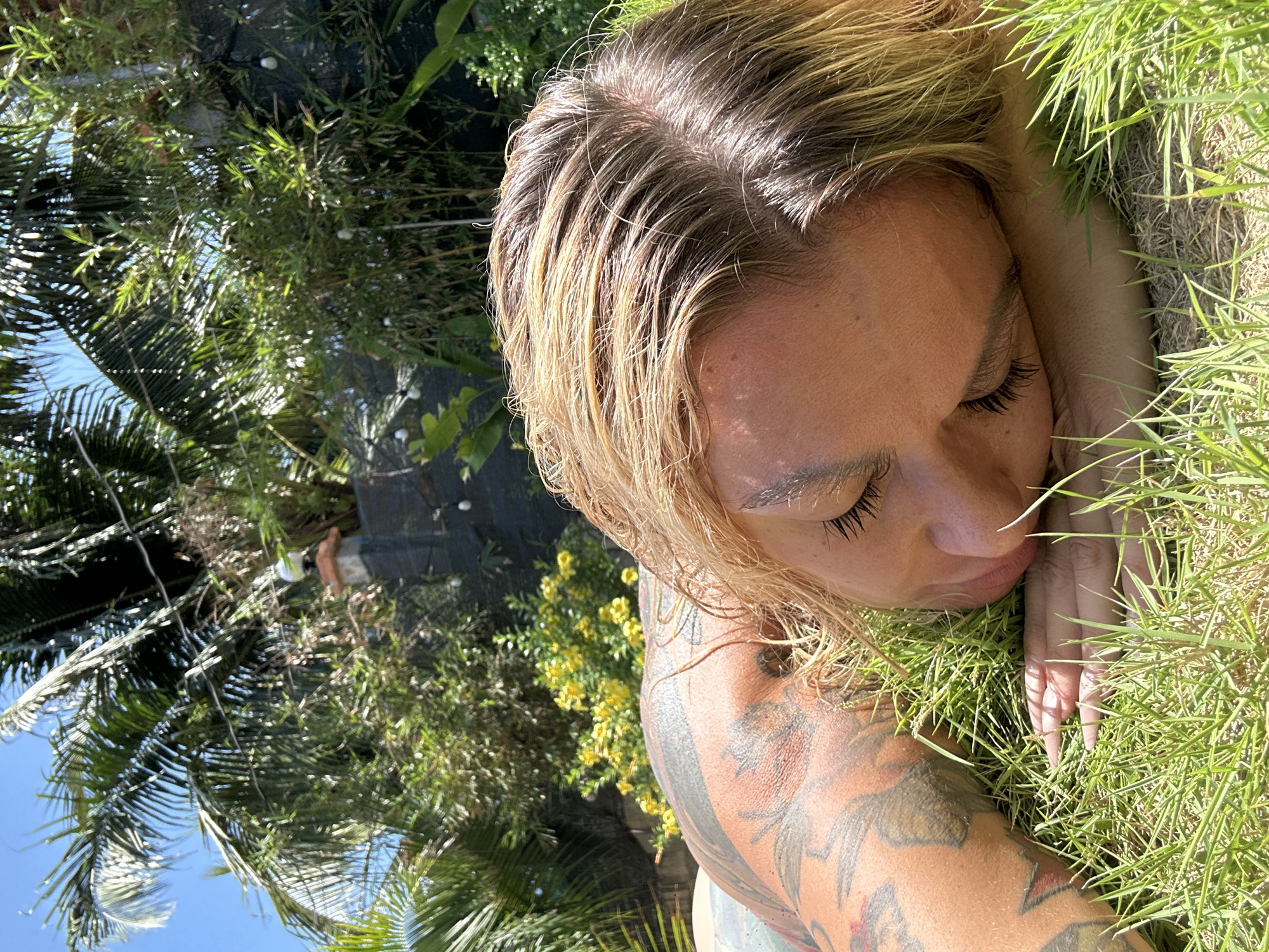 A woman with blonde, wet hair lies in the grass in Mũi Né, Vietnam. Her face is turned to the side, eyes closed, with her arm outstretched in front of her. The woman's right arm is tattooed. Behind her is a variety of foliage, including palm trees and flowering plants. The scene is outdoors under natural light.