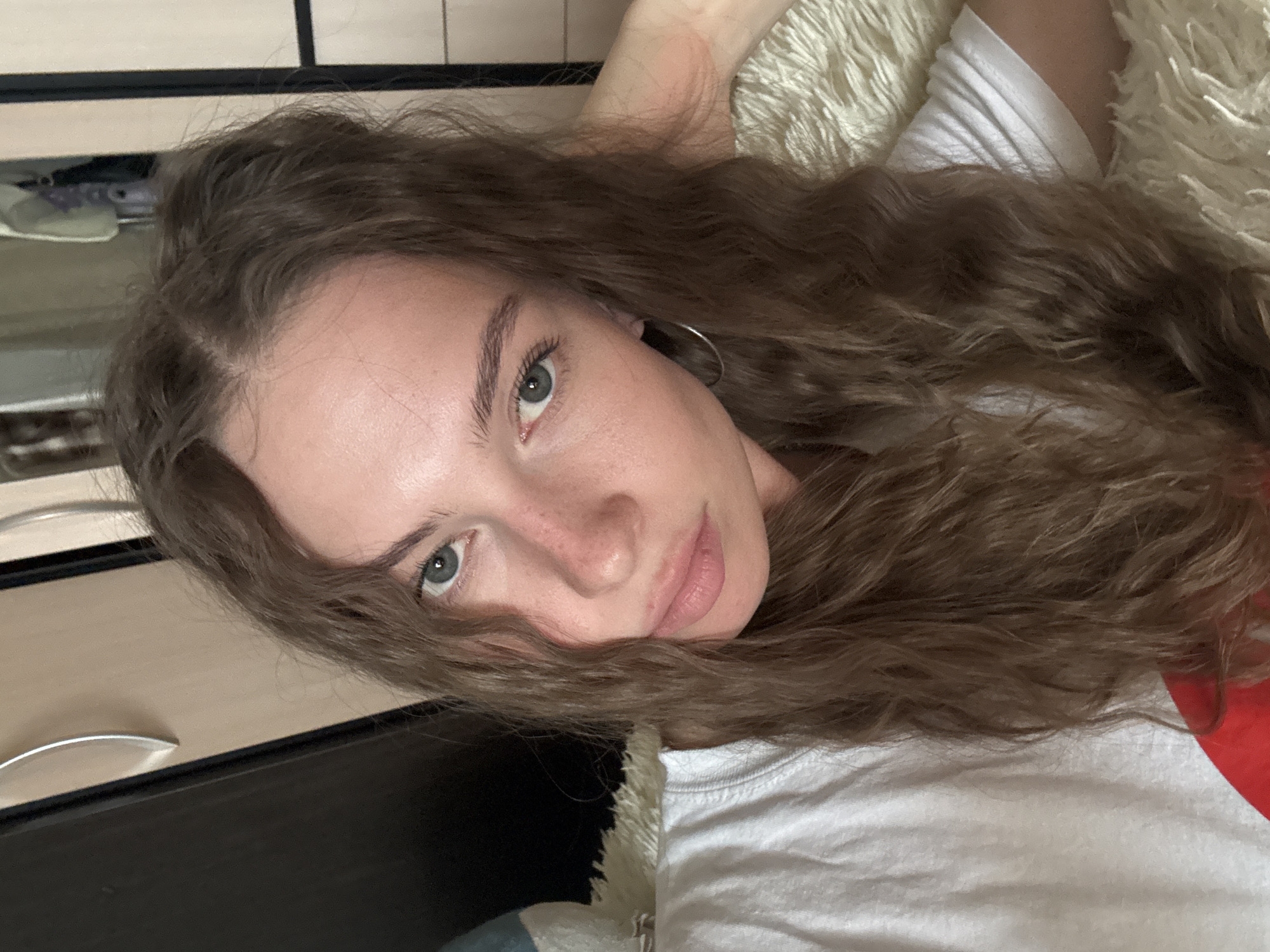 A young woman with long, wavy, light brown hair is taking a selfie in a room. She is wearing a white t-shirt and has a light tan complexion with a scattering of freckles. Her eyes are a light green-blue, and she is wearing a silver hoop earring in her left ear. Her left hand is resting on her head. The background appears to be a bedroom with a closet door visible to the left and a wardrobe to the right. A fluffy, cream-colored throw is draped over a surface behind her. The lighting suggests it is daytime, possibly indoors with natural light. The image does not contain any information that would allow for the identification of its location in the city of Perm, Russia.