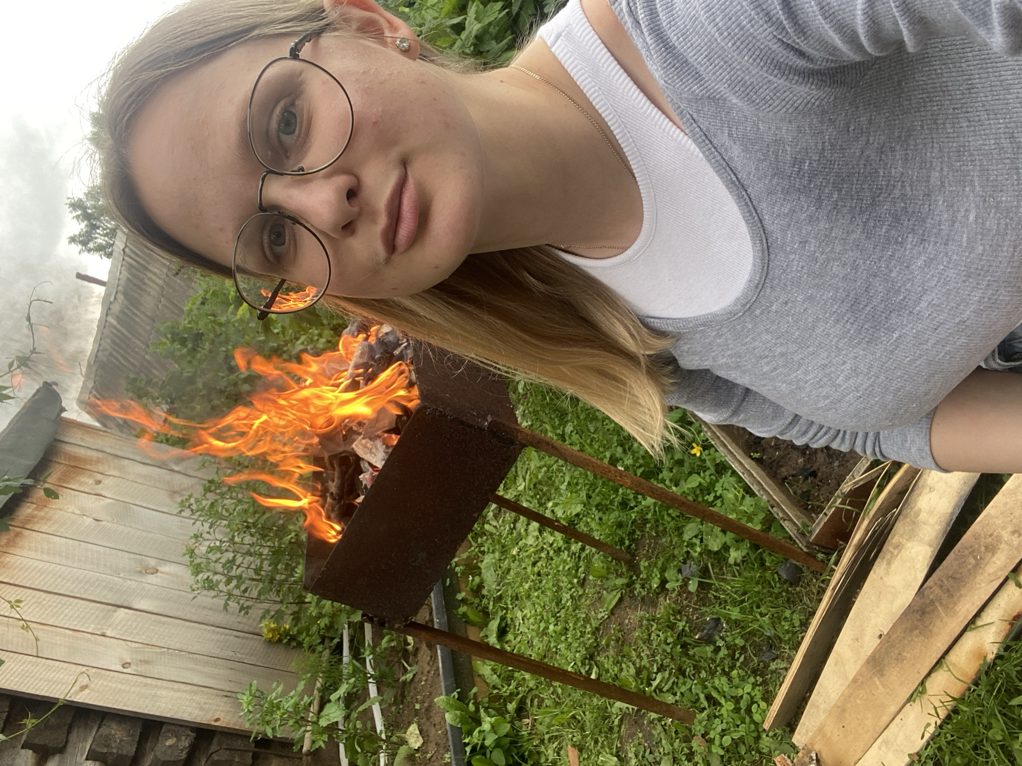 In Kauchuk Stantsiya, Russia, a young woman with blonde hair and glasses stands in a backyard setting. She is taking a selfie in front of a barbecue grill with a fire burning. She is wearing a grey long-sleeved shirt over a white tank top and a delicate necklace. The background features a wooden fence, green grass, and some plants. There are also some wooden planks in the foreground. The lighting suggests it is daytime.