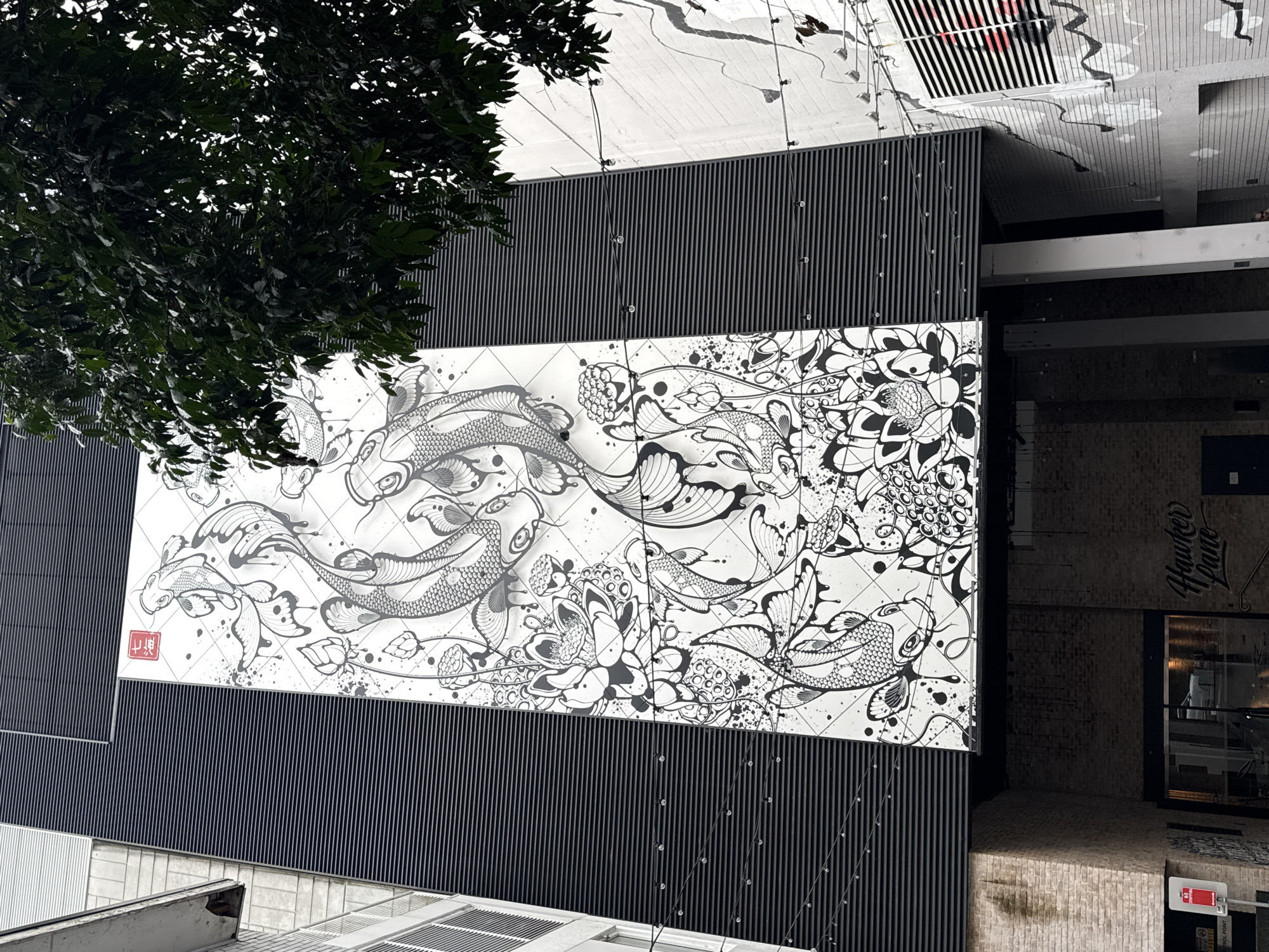 The image shows a large mural of koi fish and lotus flowers on the exterior of a building. The mural is in black and white with a red square symbol at the top left. The building is tall and has a dark, ribbed facade. A tree with dark green leaves overhangs the top right of the mural. Below the mural, there is an entrance to a building with brickwork and a sign that reads "Hawker Lane". The scene is likely in an urban environment, but the specific location in Roseville Chase, Australia is not evident from the image itself. There are no people visible in the image.