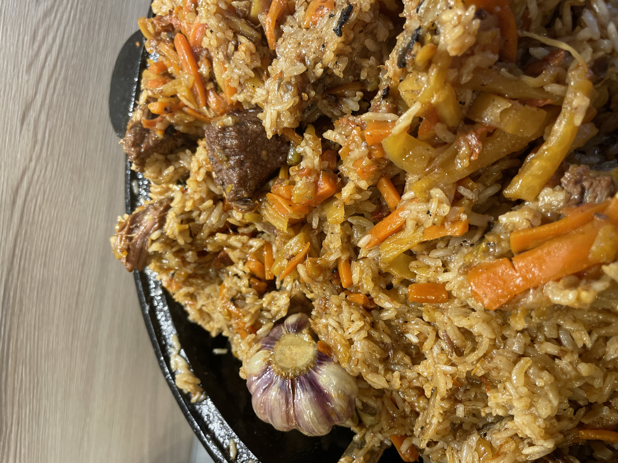A close-up shot shows a dish of pilaf, a rice-based dish, served on a black metal plate. The pilaf consists of rice, diced carrots, pieces of meat, and a whole garlic bulb. The meal sits on a light-colored wooden surface. The location is Ленинградская, Russia.