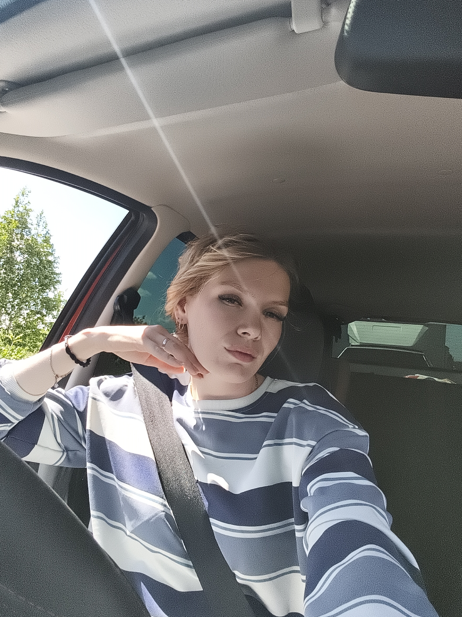 A young woman with blonde hair and makeup is taking a selfie in the driver's seat of a car, on a sunny day, with trees visible through the window. She is wearing a striped sweater and a seatbelt. Her left hand is resting on her face and her right arm is resting on her lap. The interior of the car, including the ceiling and dashboard, is visible. The location is the city of Yagul, Russia.