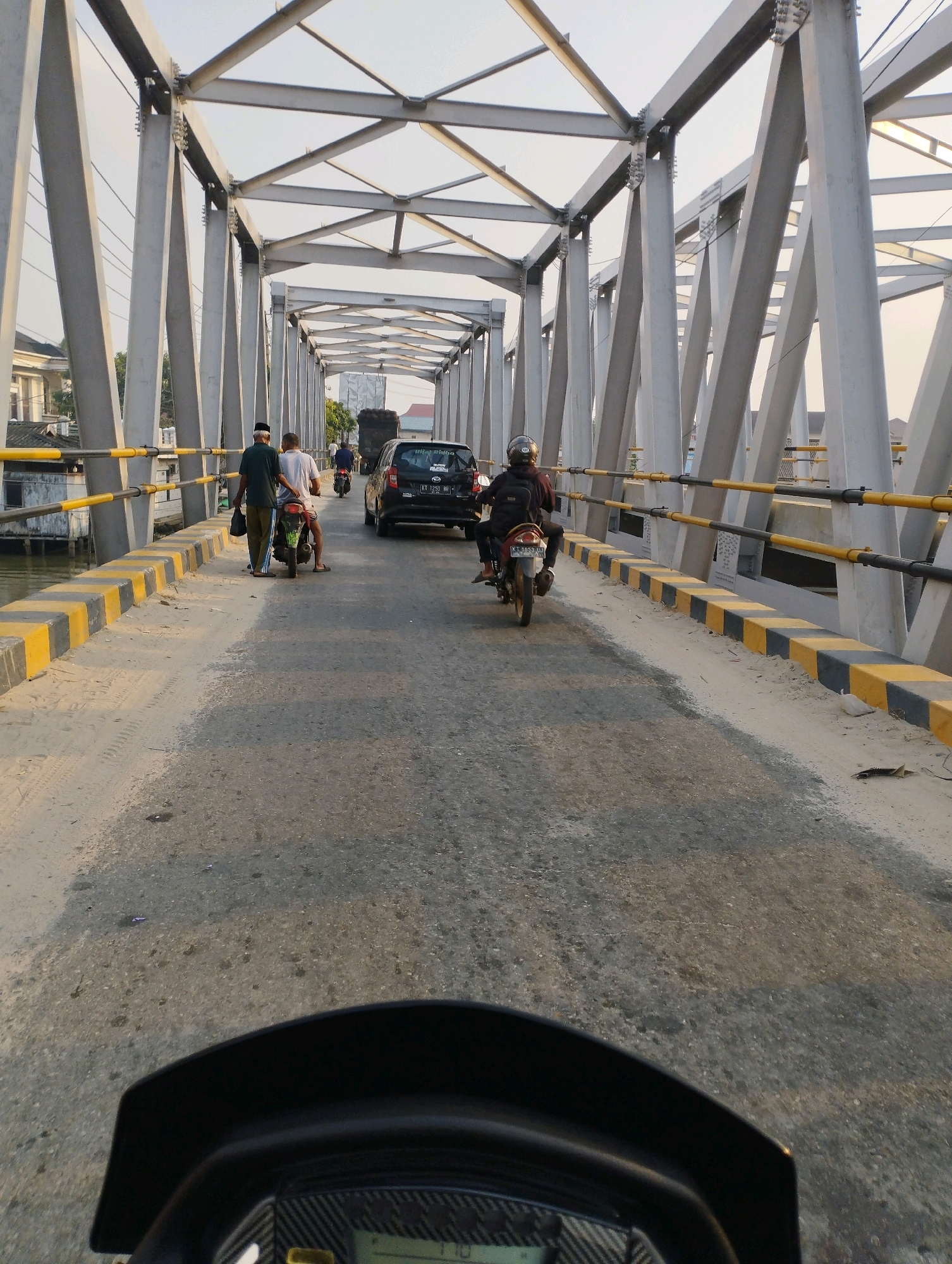 The image depicts a view from a motorcycle traveling across a bridge in Kuala Samboja, Indonesia. The bridge has a steel truss structure with gray support beams and a road surface covered in asphalt and sand. Yellow and black barriers line the sides of the bridge.
Several vehicles and people are visible on the bridge. In the foreground, the dashboard of the motorcycle the photo is taken from is visible. Further ahead, a person on a motorcycle is riding towards the viewer. To the left, two men are walking near a parked motorcycle. A black car is also on the bridge, and a truck is seen in the distance. The surrounding environment shows buildings and trees on the banks of a canal or river. The lighting suggests it is either early morning or late afternoon.