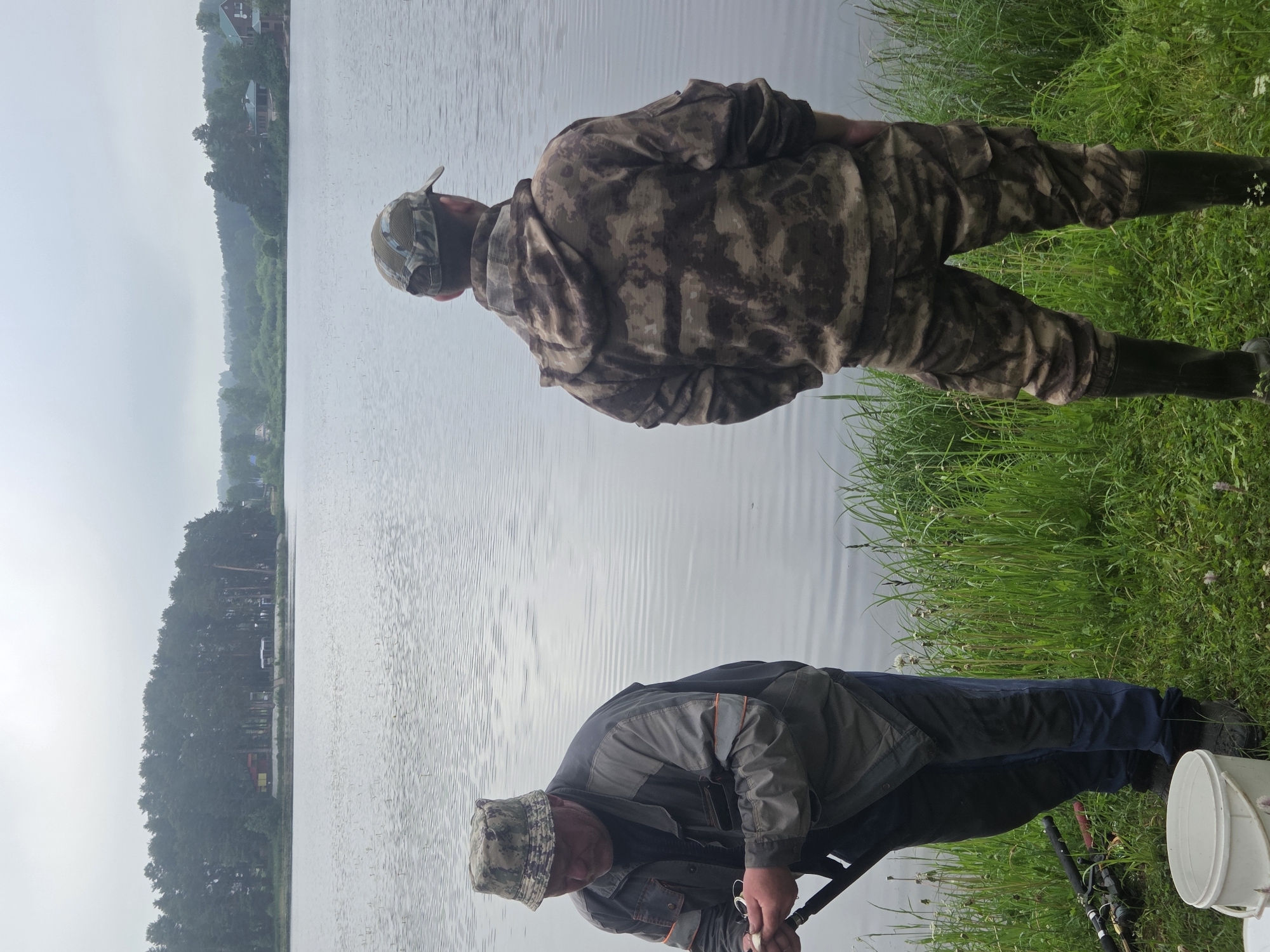 Two men are fishing on the shore of a lake in Putinovo, Russia. The man on the left is wearing a camouflage bucket hat, a grey and black jacket, and dark pants. He is holding a fishing rod and appears to be baiting a hook. The man on the right is wearing a camouflage baseball cap and a camouflage hooded jacket with matching pants. He is standing with his back to the camera, looking out at the lake. Both men are wearing dark rubber boots. The lake is calm and reflects the overcast sky. The shore is covered in green grass and some weeds. In the background, across the lake, there is a dense line of pine trees, and further in the distance, there are some buildings and houses visible. A white bucket and fishing rods are placed on the grass near the man on the left. The overall atmosphere is peaceful and rural.