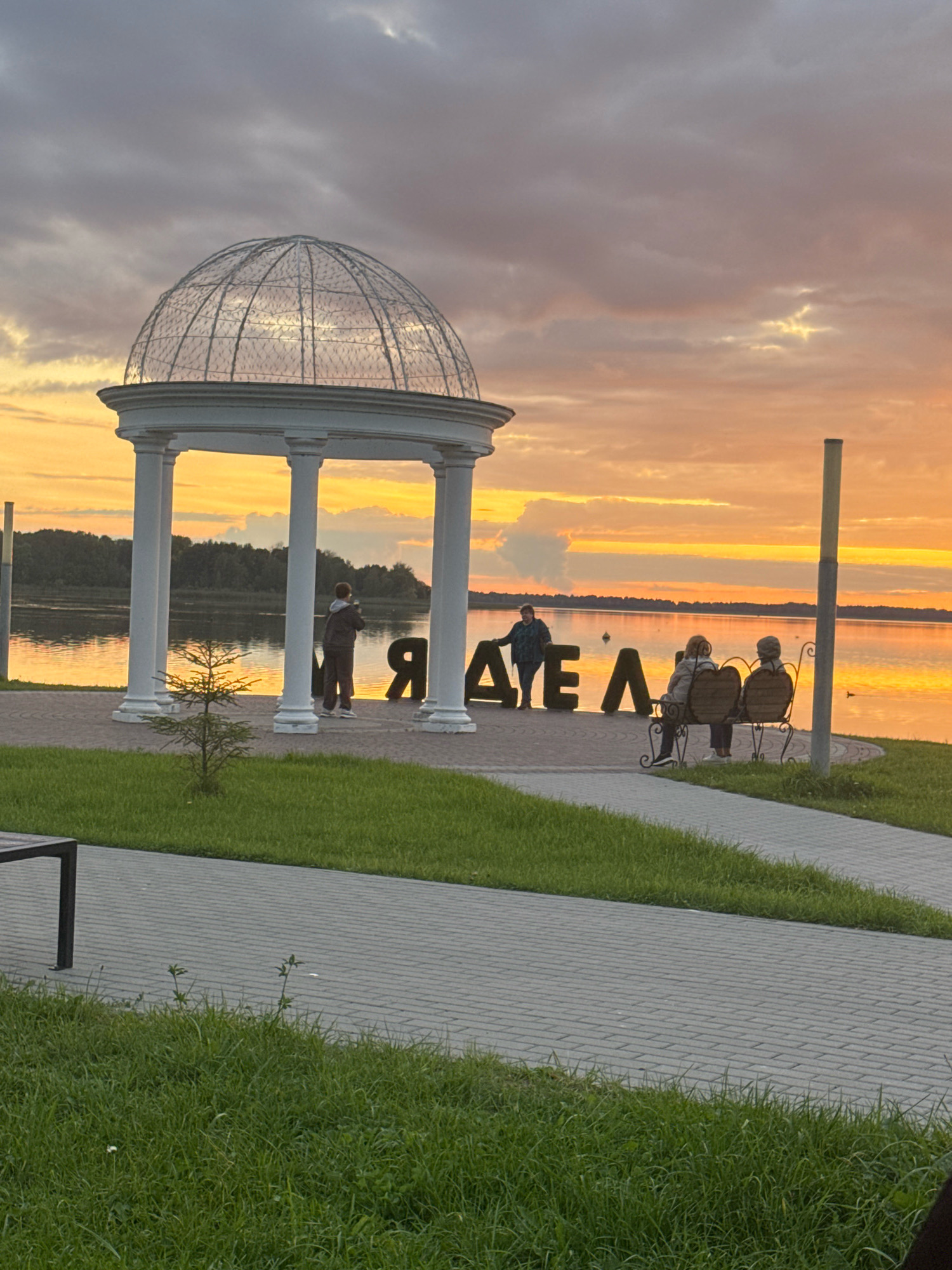In Myadzyel, Belarus, a scenic view captures a sunset over a body of water. In the foreground, a paved walkway and grassy area lead to a white gazebo with a domed roof supported by columns. Large, dark letters spelling "ЯДЗЕЛ" (Myadzyel) are placed near the gazebo. Two individuals are standing near the letters, one seemingly taking a photograph. Further to the right, three people are seated on a bench, facing the water. The sunset casts a warm orange glow across the sky and reflects on the calm water. Trees line the far bank of the water. The scene is tranquil, with the natural beauty of the sunset and the architectural elements of the gazebo and lettering creating a picturesque setting.