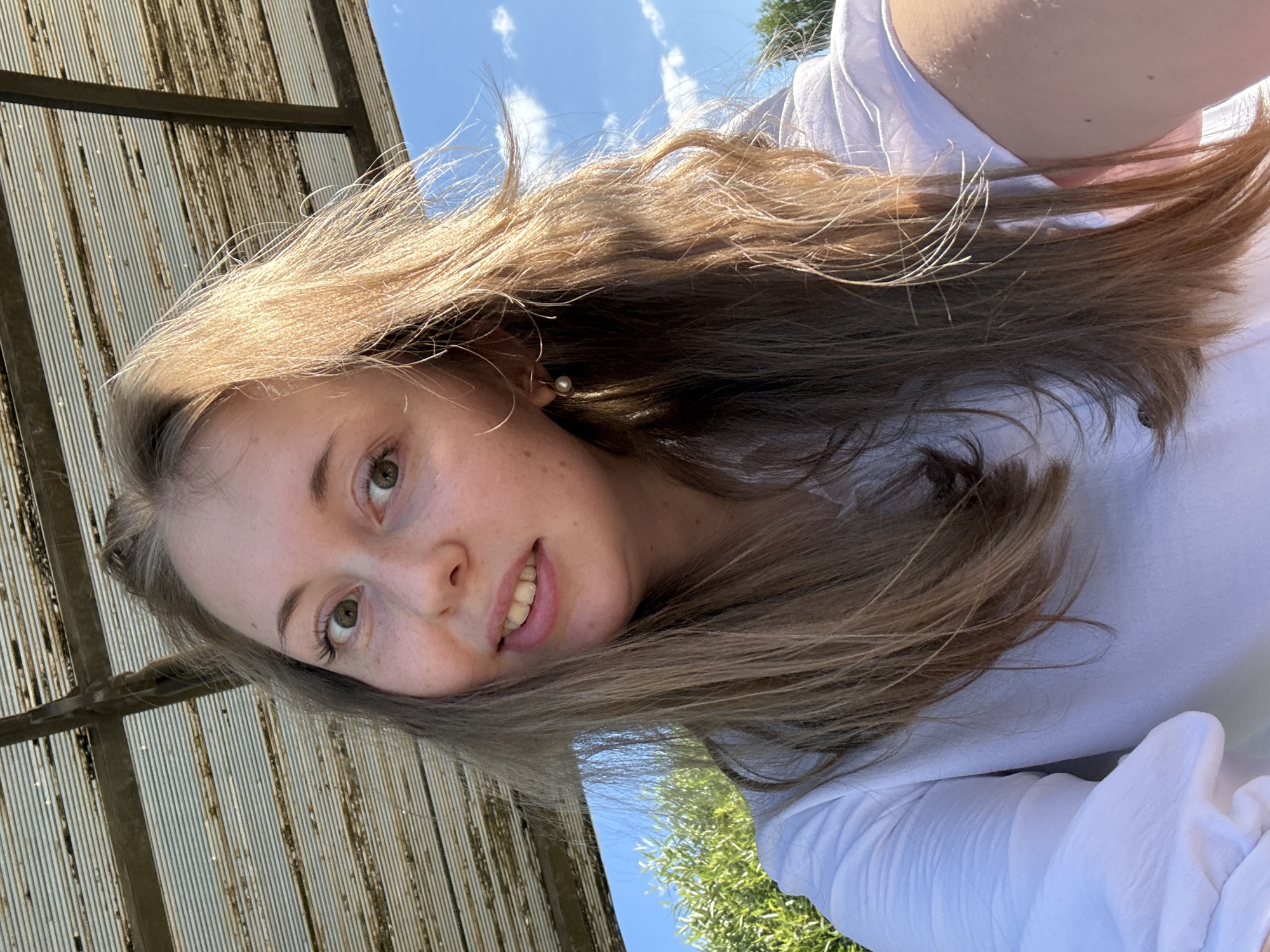 A young woman with long, light brown hair is taking a selfie outdoors. She is wearing a white shirt and a pearl earring. The background shows a blue sky with clouds, green foliage, and a corrugated metal awning. The lighting suggests it is daytime and sunny.