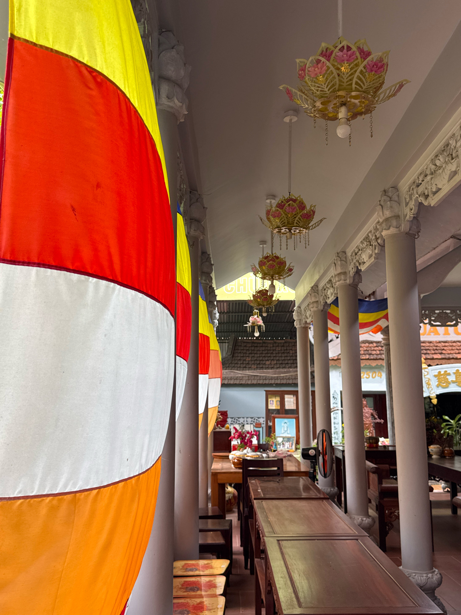 The image shows an outdoor covered walkway at what appears to be a Buddhist temple or cultural site in Nha Trang, Vietnam. Prominently featured in the foreground are large, vertical Buddhist flags with distinct bands of yellow, red, and white, partially obscuring the view beyond. The walkway is supported by white, fluted columns adorned with intricate carvings. Hanging from the ceiling are decorative lotus-shaped lanterns. In the background, visible through the walkway's columns, are more Buddhist flags, tables and chairs set up, and a glimpse of traditional tiled roofs. A black oscillating fan is mounted on one of the columns. The scene suggests a tranquil, cultural, and religious setting within the city.