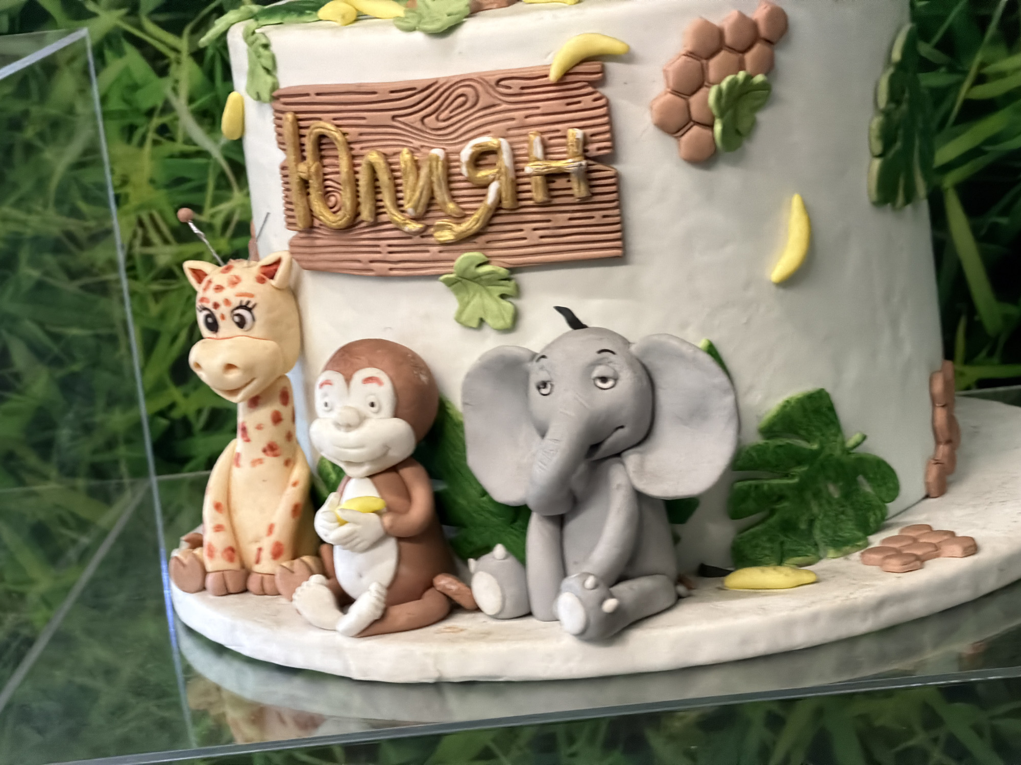 A close-up shows a decorated cake. The cake features a giraffe, monkey, and elephant made of fondant. A wooden-style sign with a name in gold lettering is placed on the side. Green leaves and bananas also decorate the cake, as well as a design resembling honeycomb. The cake sits on a clear surface, possibly a display case, and the background includes blurred greenery. The location is unknown, there is no indication of Sofia, Bulgaria.