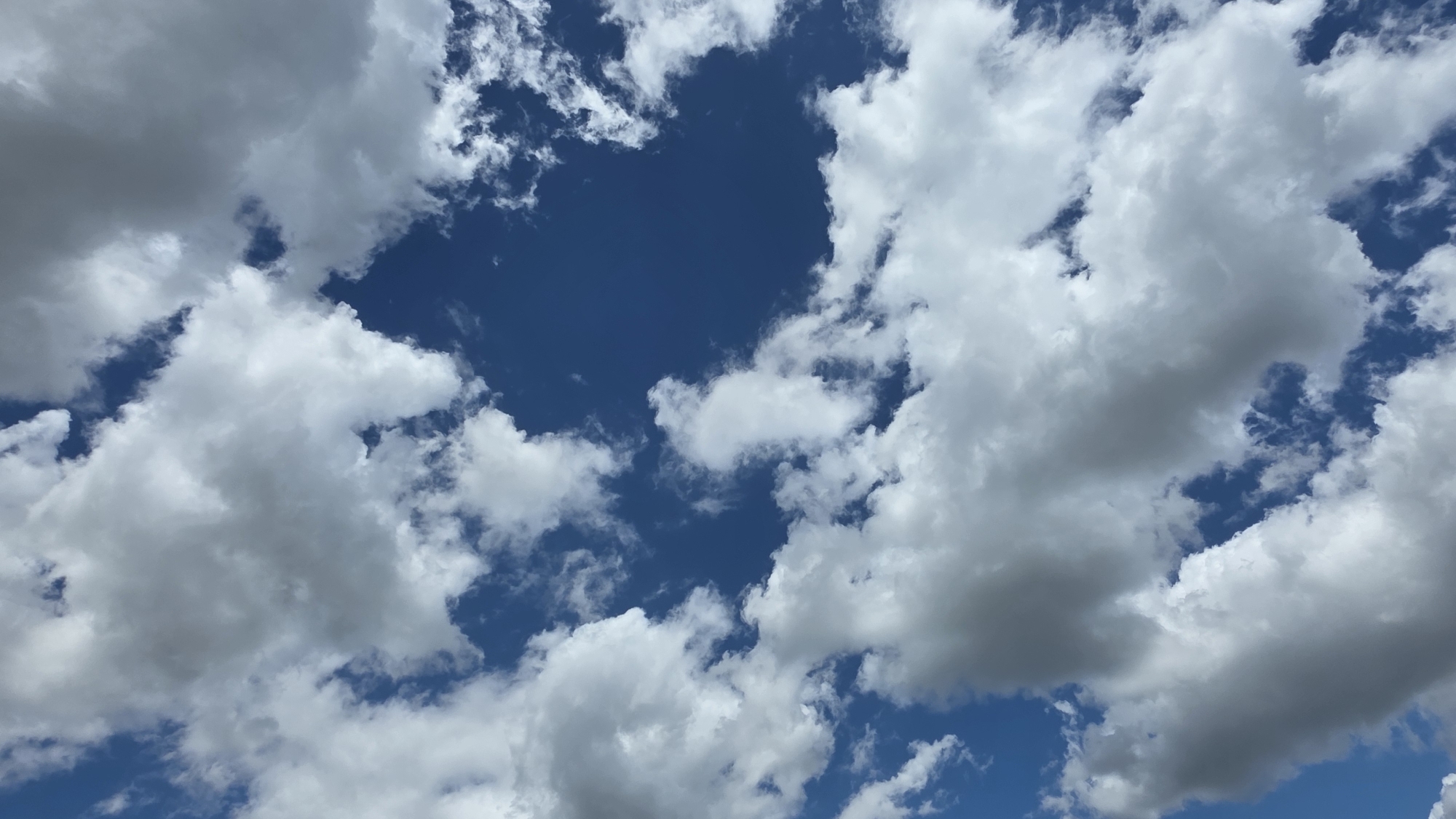 A wide shot captures a blue sky filled with white and gray clouds. The clouds are scattered and varied in size and density, with some appearing fluffy and bright, while others are darker and more ominous. Patches of clear blue sky are visible between the clouds, creating a dynamic and textured appearance. The image is taken from a low angle, looking up towards the sky, suggesting a perspective from the ground. The lighting indicates it is daytime, likely with the sun obscured by some of the clouds, casting shadows and creating depth. There are no people, structures, or other objects visible in the frame. The scene is purely natural, focusing on the atmospheric conditions of the sky.