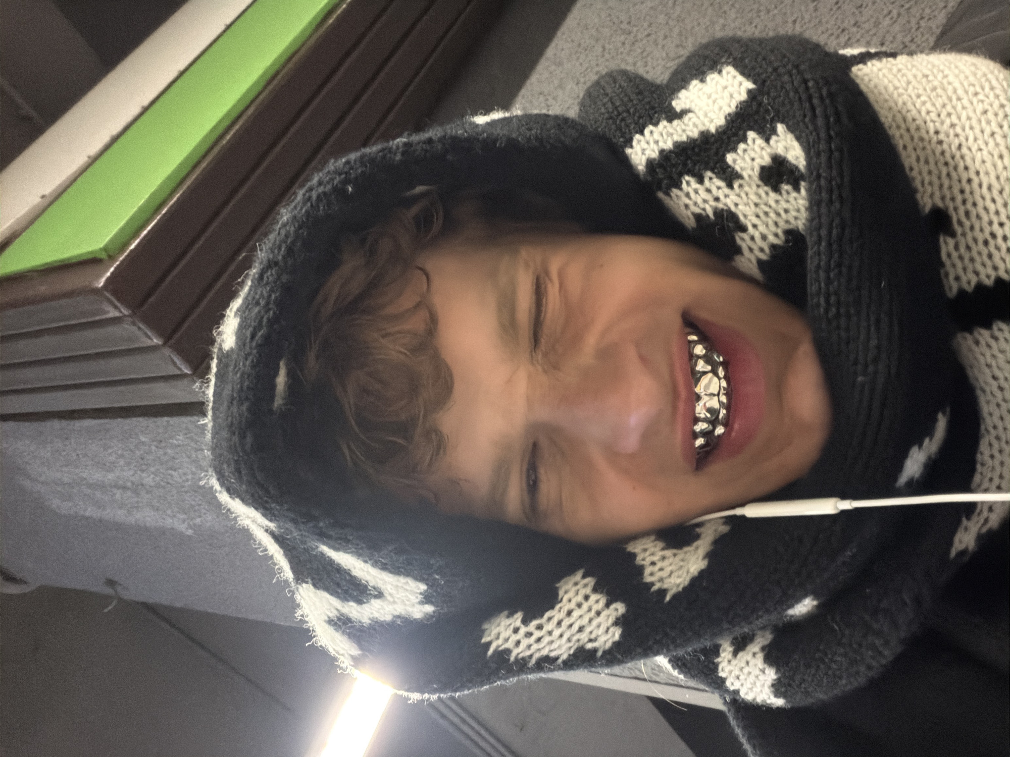An indoor portrait, rotated 90 degrees clockwise, features a young person with curly brown hair, wearing a black and white knitted hooded garment. The garment has a distinct pattern of jagged lines or abstract shapes. The person is smiling broadly, their eyes squinted or closed in a strong expression, possibly joy or playfulness. They prominently display silver grillz on their upper teeth, reflecting light. A white earbud wire is visible, extending from their ear area down across their shoulder.
The background reveals interior architectural elements. Diagonally behind and above the person's head are brown rectangular panels or tiles, alongside a bright lime green strip and a lighter beige/off-white strip. Below the person, a grey textured surface and a bright artificial light source are visible, suggesting a modern or urban interior setting within Milan, Italy. The lighting is artificial, illuminating the subject and the immediate surroundings. No text is visible in the image.
