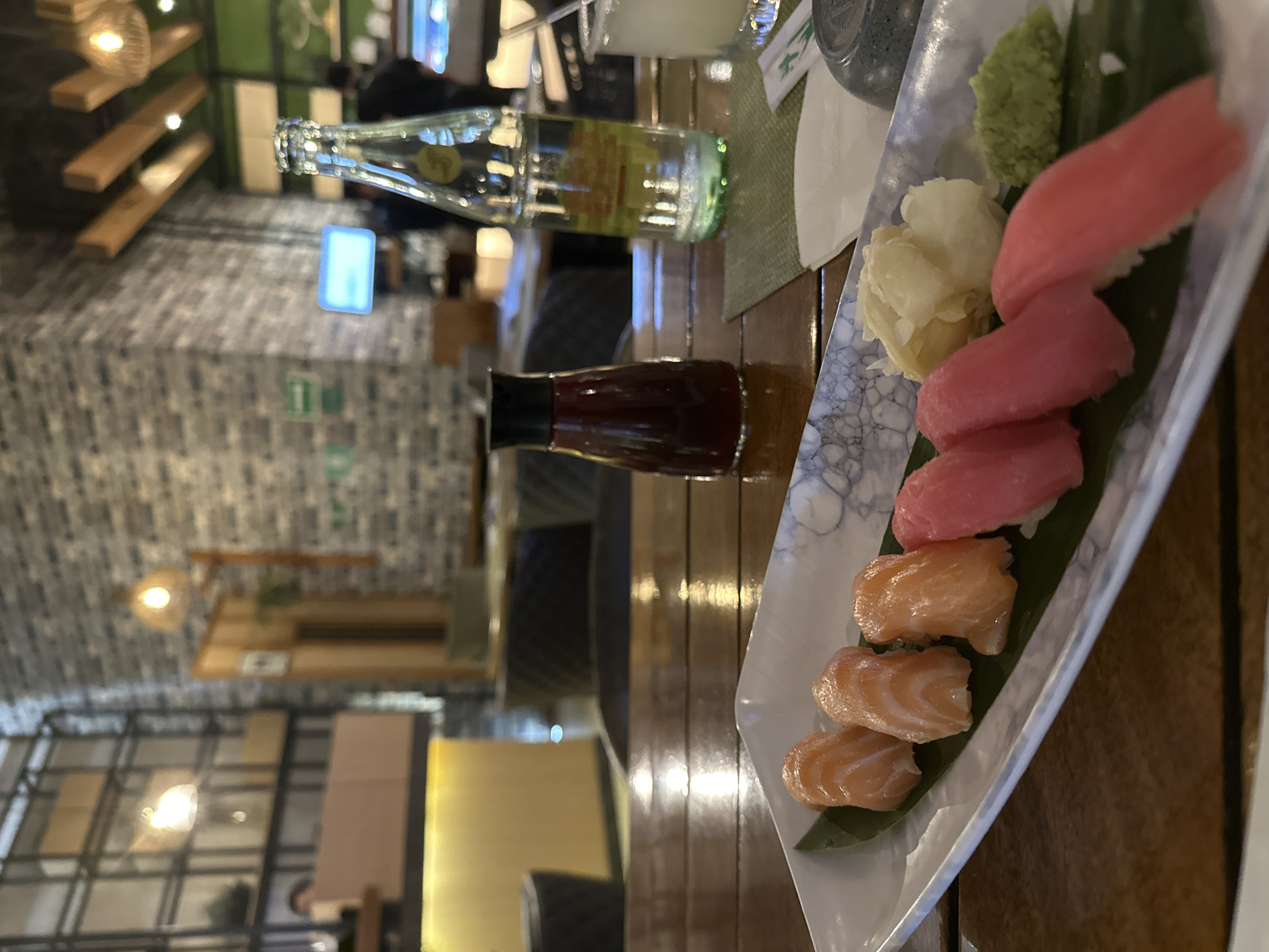 The image shows a plate of sushi on a wooden table. The sushi consists of salmon and tuna nigiri, served with wasabi and ginger. Also visible is a bottle of water and soy sauce. The location is San Bernardino Tlaxcalancingo, Mexico. The environment suggests a restaurant setting. No people are clearly visible, although there are chairs.