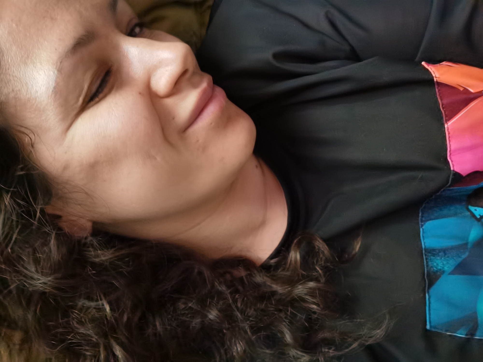 A close-up, high-angle shot shows a person lying down, with their head resting on a surface covered by a dark fabric. The person has dark, curly hair and their face is partially visible, with their eyes closed and lips slightly parted. The person is wearing a dark t-shirt with a colorful abstract design on the right side. The image appears to be taken indoors. There is no information to suggest this location is in the city of Chaikovsky, Russia.