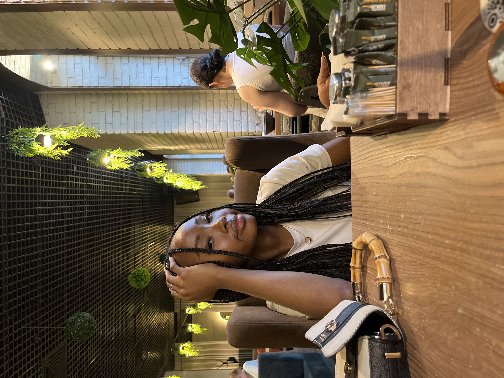 The image depicts a young Black woman with long, braided hair, wearing a white t-shirt and a necklace, seated at a wooden table in what appears to be a restaurant. She is resting her head on her hand and looking towards the camera. In the foreground, a handbag with a bamboo handle and a striped detail is visible on the table. To the right, a wooden tray holds an assortment of individually packaged sugar, salt, and pepper packets, along with toothpicks. In the background, a woman with her hair in a bun is seated at another table, facing away from the camera. The restaurant's interior features a black grid ceiling with hanging greenery and industrial-style lighting, and exposed brick walls. The overall atmosphere suggests a casual dining setting.