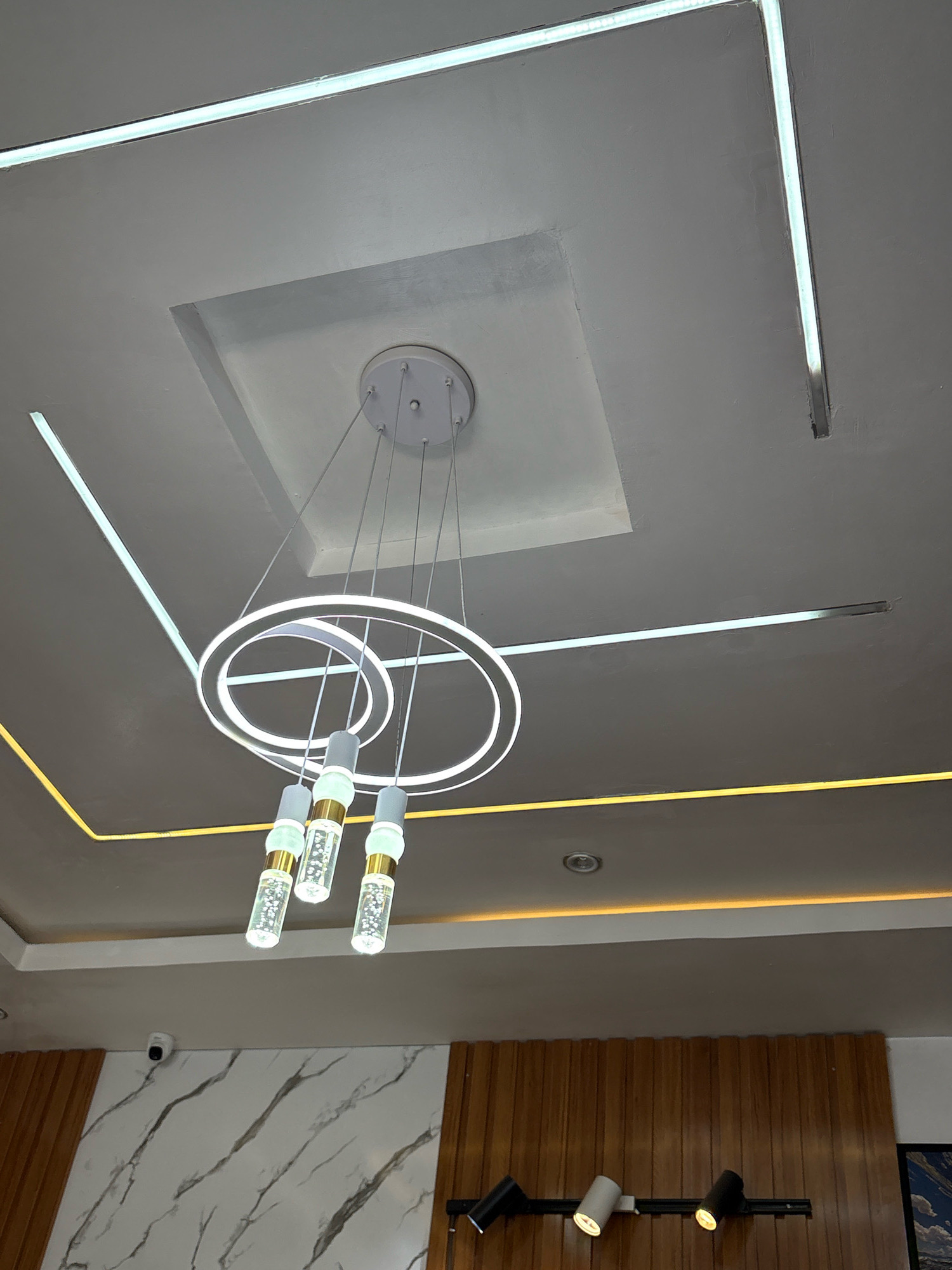 The image captures the interior of a modern ceiling with multiple light fixtures. A prominent contemporary chandelier with concentric circular elements and three hanging cylindrical glass lights is centered. Linear LED light strips are embedded in the ceiling, forming geometric patterns, with a subtle yellow accent strip along a recessed area. The ceiling itself is a multi-level design, with the central chandelier suspended from a recessed square panel. In the background, a wall features a marble-like pattern and vertical wooden paneling. There are also track lighting fixtures visible on the wooden paneling, and a small white security camera mounted on the wall. The overall scene suggests a well-lit, contemporary indoor space, possibly a commercial establishment or a modern residence. There are no people or explicit actions depicted in the image. The location in Jos, Nigeria, cannot be determined from the visual information alone.