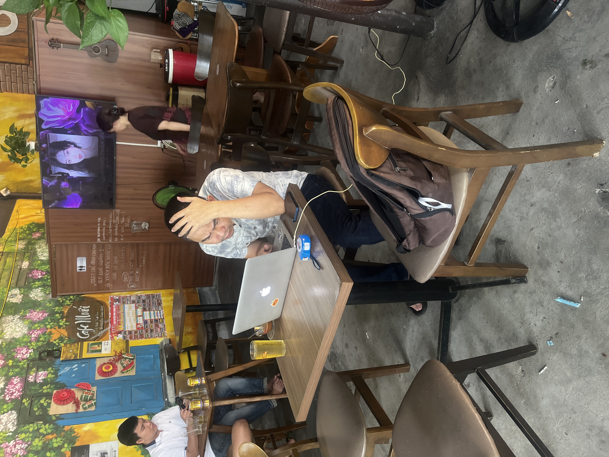 A man is seated at a table in a cafe in Da Nang, Vietnam, working on a laptop. He appears stressed, with his hand on his forehead. A glass of beer and a measuring tape are on the table. The cafe has a colorful mural on the wall and a television playing a video of a woman. A woman in the background is walking away from the camera. Other patrons are visible at tables in the background. The floor is concrete, and there are several chairs and tables.
