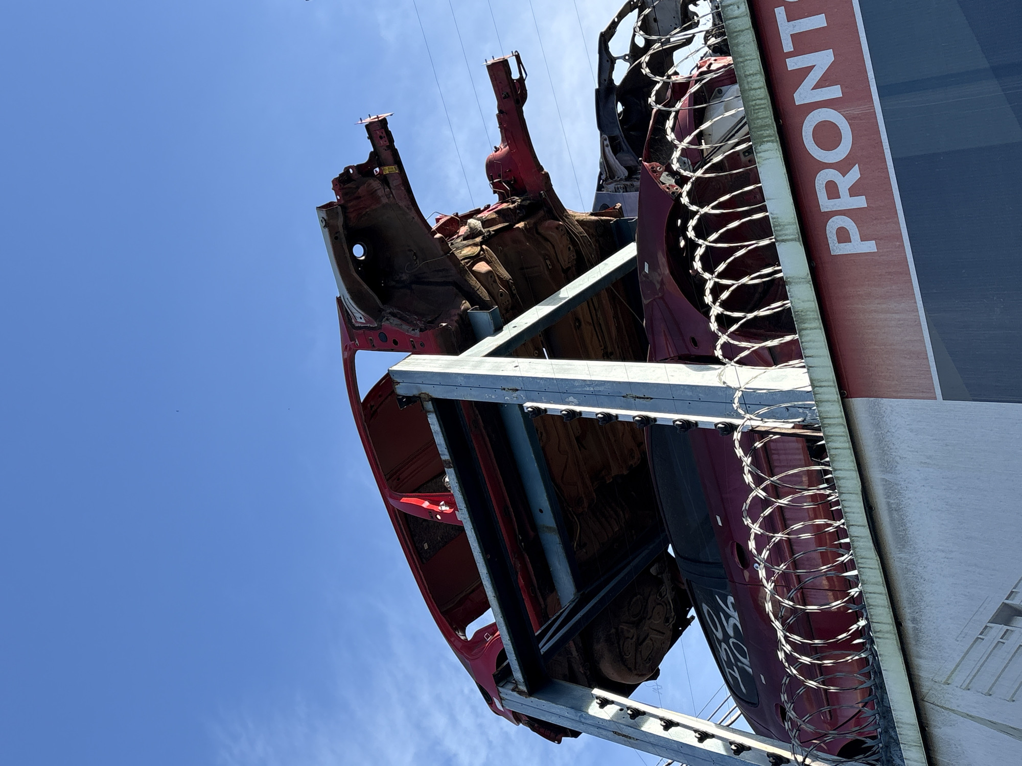 The image shows a car graveyard under a clear blue sky. Several car shells, mostly red, are stacked on a metal rack. Some of the car shells are partially dismantled, exposing their internal structures. In the foreground, a wall topped with barbed wire separates the car graveyard from a structure with a large red sign that reads "PRONTO" in white lettering. The scene appears to be an outdoor salvage yard or recycling facility, likely located in a commercial or industrial area of São Paulo, Brazil, given the presence of power lines and the overall urban-industrial aesthetic. There are no people visible in the image.