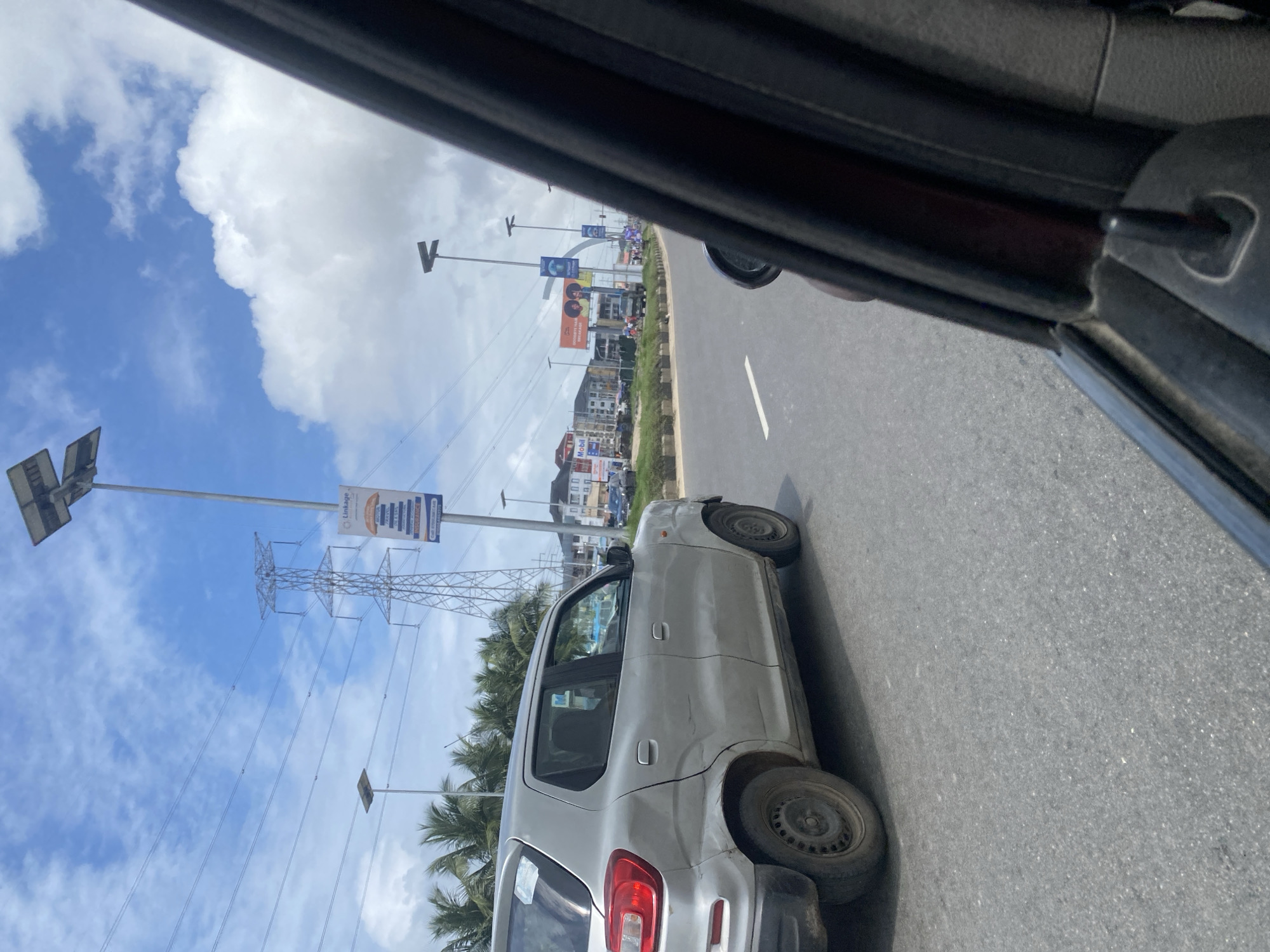 The image captures a street view from a moving vehicle in Lekki Peninsula II, Nigeria, under a partly cloudy sky. A silver car is prominently featured on the asphalt road, showing visible dents and scratches on its side. In the background, palm trees line the median, and various buildings, including what appear to be residential or commercial structures, are visible. A tall electricity pylon with power lines stretches across the sky, alongside streetlights with solar panels. Several billboards, including one for "Mobil," are also present. The scene depicts a typical urban or suburban street in the area.