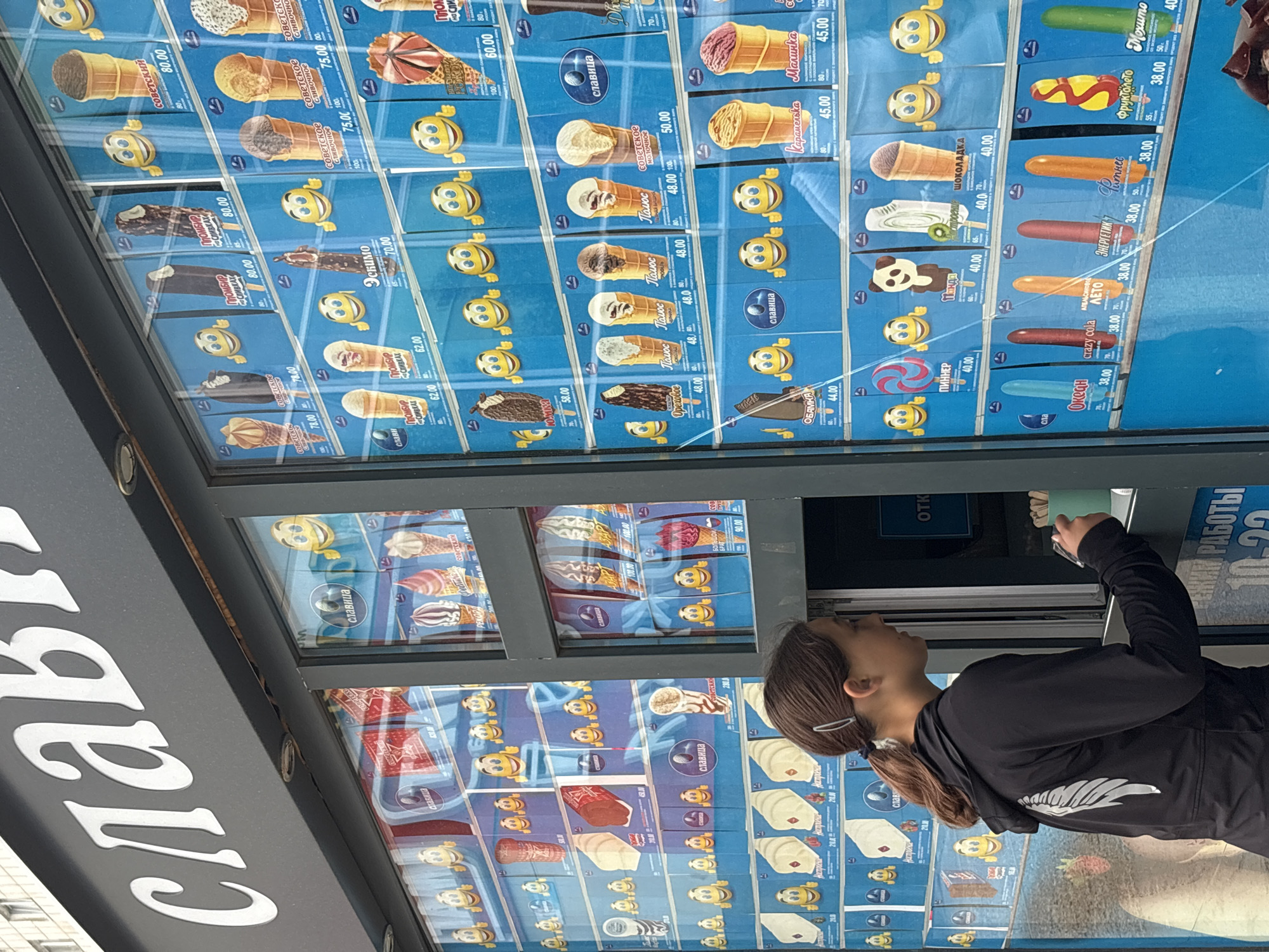 A young person with brown hair in a ponytail is standing in front of a glass display case of ice cream at an outdoor kiosk. The kiosk, located in Chaikovsky, Russia, has a gray exterior and the word "Славiцa" written in large white Cyrillic letters. Inside the display case are numerous ice cream options, each with a picture, a price, and a small cartoon smiley face. The prices are labeled in Russian currency. The person is reaching towards the service window of the kiosk. The sign on the kiosk indicates it operates from 10:00 to 22:00.