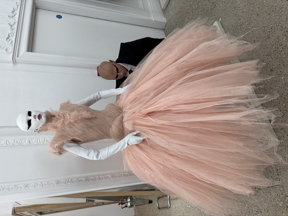 A light pink tulle gown engulfs the frame, worn by a mannequin with white face paint and dramatic black eyeliner. White gloves extend up the mannequin's arms. Behind the mannequin, a bald figure in a black suit and white shirt peers out. The background includes a white wall with ornate molding, a door, and a concrete floor. Part of a metal structure is visible at the bottom of the image.
