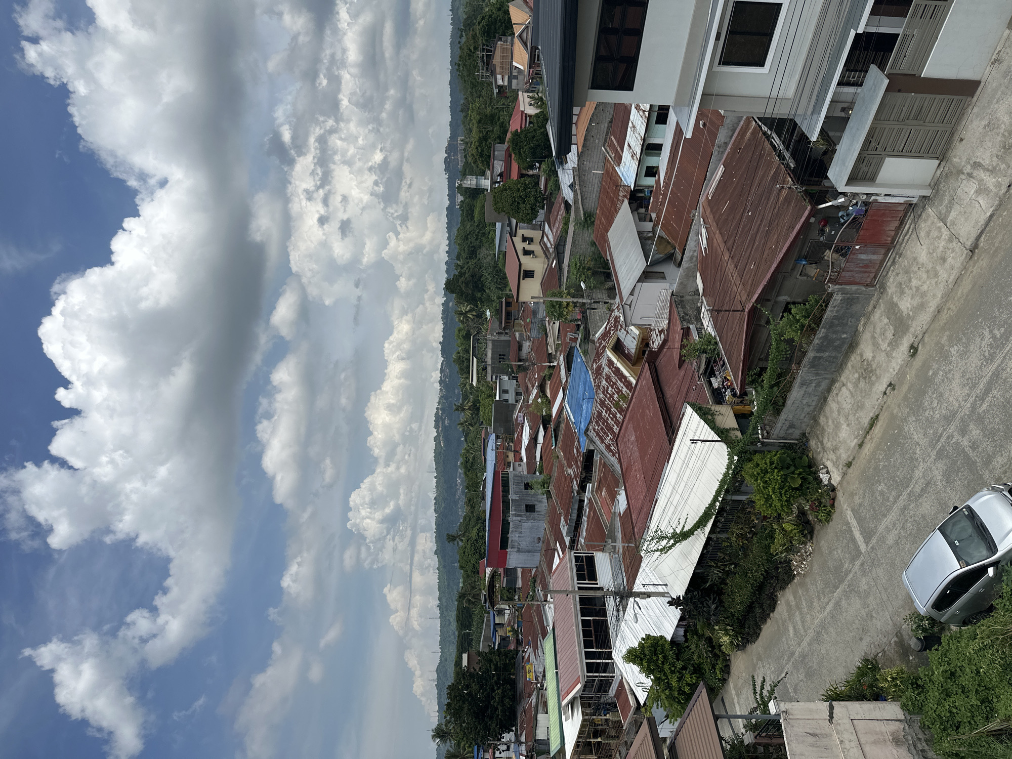 An elevated view shows a street scene in Davao City, Philippines, with a mix of residential and commercial buildings. The structures are mostly made of concrete and corrugated metal roofing in various colors. Vegetation is interspersed throughout the neighborhood. A silver car is parked on the street, and the sky above is filled with scattered clouds.