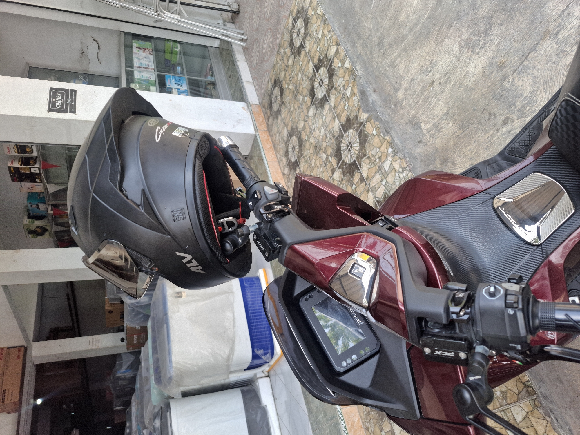 A close-up, eye-level, outdoor, medium shot captures a red scooter parked on a tiled pathway in front of a store. A black motorcycle helmet is attached to the handlebars, facing forward, with its visor up. The scooter's digital dashboard is visible in the foreground, displaying a screen with a reflection of trees and sky. The store behind the scooter has shelves stocked with various products, including what appear to be small appliances and packaged goods. The pathway is adorned with a patterned tile design, and a concrete sidewalk runs alongside it. The overall scene suggests a common urban setting in Banjarmadu, Indonesia, likely during daylight hours.