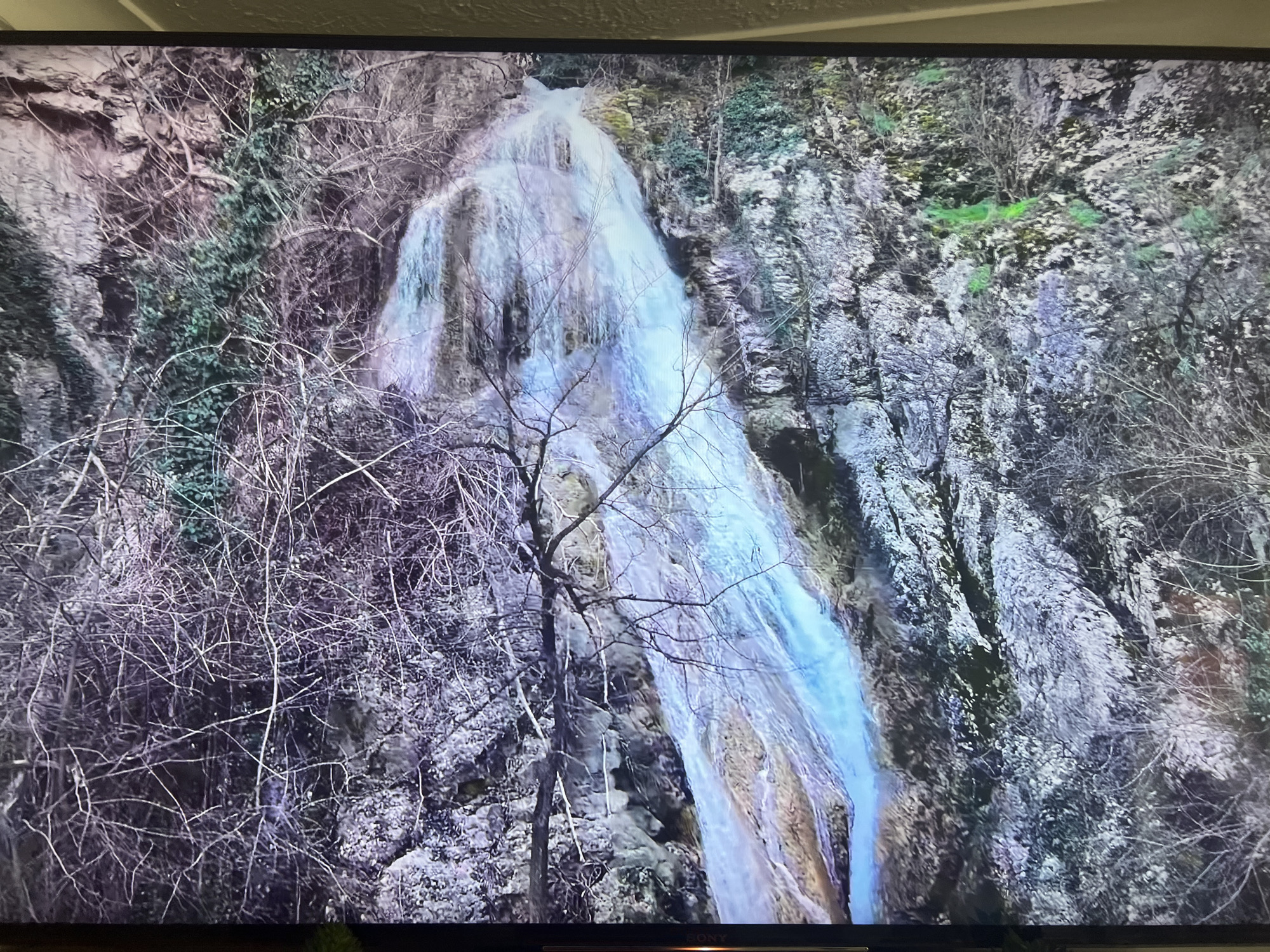 A waterfall cascades down a rocky cliff face, partially obscured by bare trees and foliage. The waterfall appears as a blurred white and blue stream against the darker rock. The surrounding vegetation is predominantly leafless, suggesting winter or early spring. Patches of green growth can be seen on the cliff face. The scene is likely a natural landscape, though the overall image has been displayed on a screen. The image does not contain any people. There are no recognizable interactions or actions. The location is not discernible from the image alone. Further information would be needed to locate the scene in Basingstoke and Deane, United Kingdom.