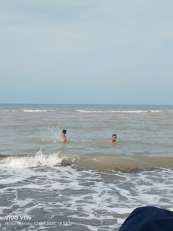 This image captures two individuals partially submerged in murky, grey-brown ocean water under a light blue to grey sky. The scene is located on a beach in Limbangan, Indonesia. Waves with white foam are visible in the foreground and midground.
On the left, one individual, an adult male or adolescent with dark hair, is seen from the back, chest-deep in the water, with water splashing around their shoulders. On the right, a second individual, an adult male or adolescent, is chest-deep in the water, facing towards the viewer with a neutral facial expression. Both individuals are shirtless. A dark, possibly navy-blue fabric object is present in the bottom right foreground, partially obscuring the water. A digital timestamp "vivo Y19s Akhtar & Arvan, 12 Okt 2025, 14.53" is overlaid in the bottom left corner.