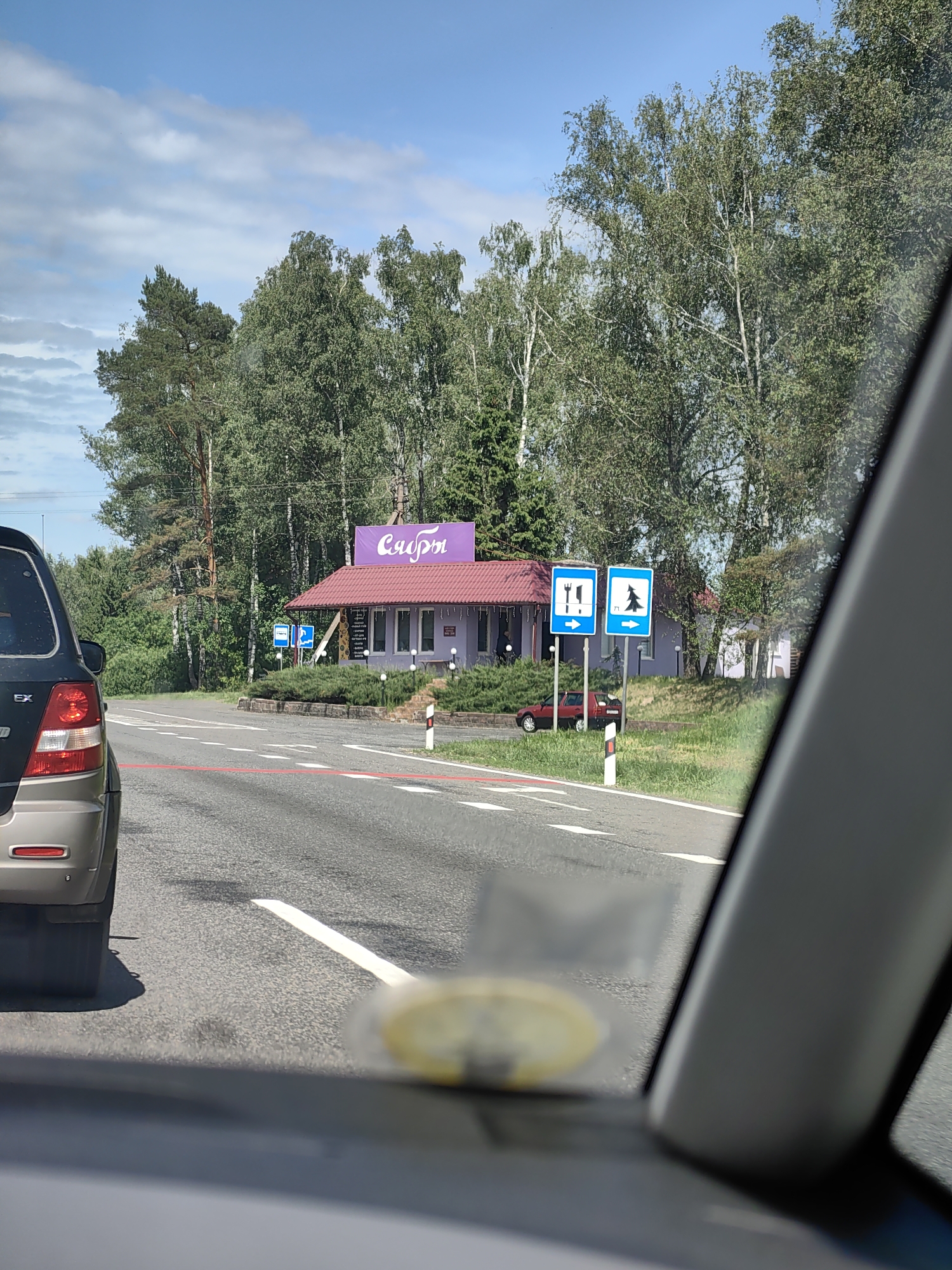 The image captures a roadside scene on a partly cloudy day in Бараўскі сельскі Савет, Belarus, viewed from inside a vehicle. In the foreground, the rear of a dark gray or brown SUV, possibly a Kia Sorento (indicated by visible "EX" badging), is partially visible on the left. A prominent red horizontal line is painted across the asphalt road, functioning as a stop line or demarcation for an intersection or crossing.
To the right of the road, a single-story building with light purple exterior walls and a red tiled roof stands behind a small landscaped area with trimmed green bushes. A large purple sign with the word "Сябры" (Syabry, meaning "Friends" in Belarusian) in white Cyrillic script is mounted on the roofline of the building. In front of the building, two blue square informational road signs are erected on poles: the left sign displays a fork and knife pictogram, indicating a restaurant or food service, and the right sign displays a pine tree pictogram, signifying a recreational or forest area, both accompanied by right-pointing arrows. A small, dark red sedan is partially visible, parked beyond the delineator post and partially obscured by the building and grass. A white and red striped delineator post is positioned on the road's shoulder.
The background is dominated by a dense line of trees, including both light-barked deciduous trees (likely birches) and darker conifers (likely pines), extending across the horizon. The sky is blue with scattered white cumulus clouds.