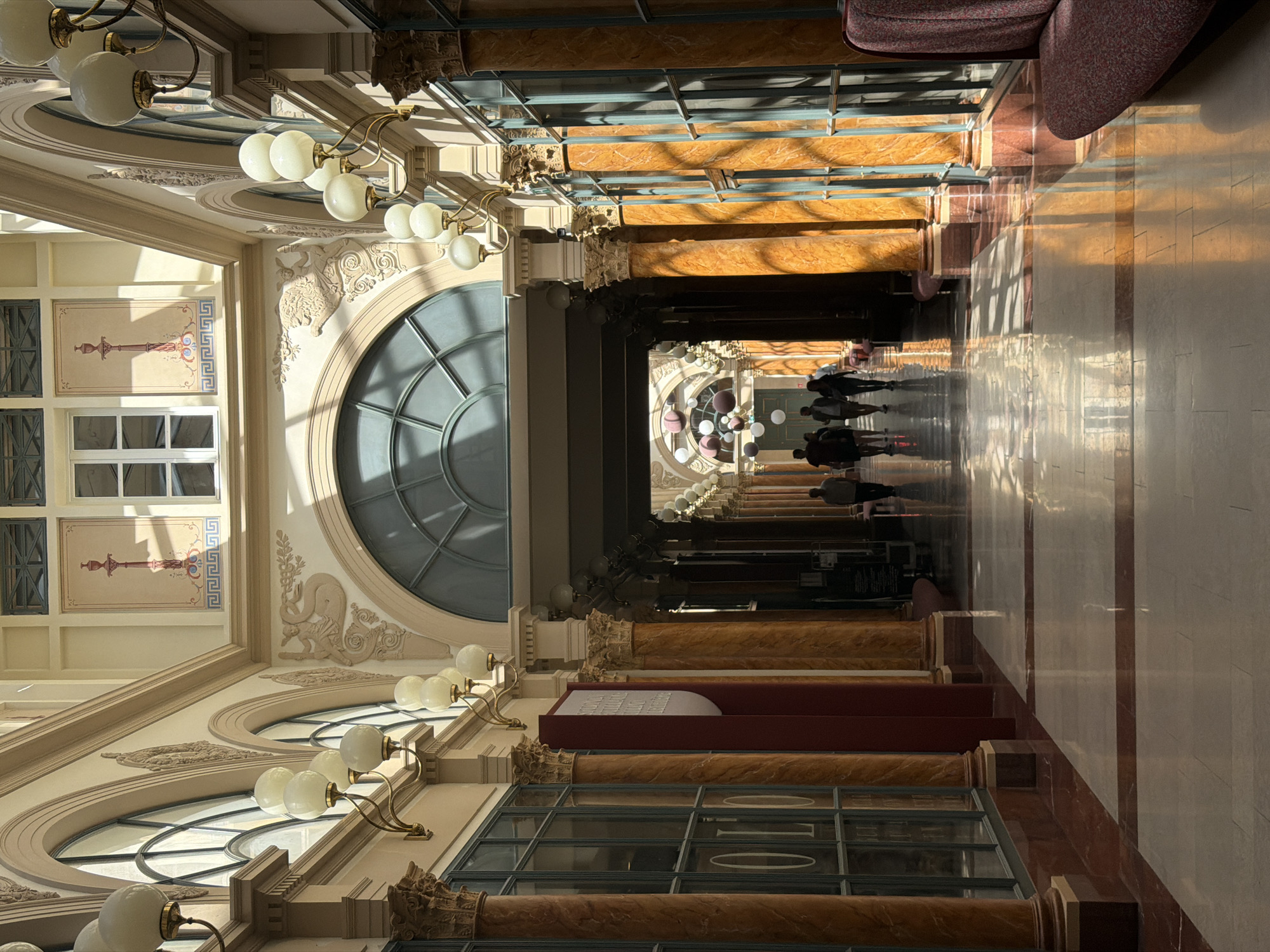 The image depicts a corridor in the Galerie Vivienne, a historical covered passage in Paris, France. The corridor is lined with marble columns and arched windows, creating a perspective view. Light fixtures are visible along the ceiling, and the floor is tiled with geometric patterns. People are walking in the distance, suggesting movement within the space. The architecture is ornate, showcasing intricate details and decorative elements. The environment appears to be indoors.