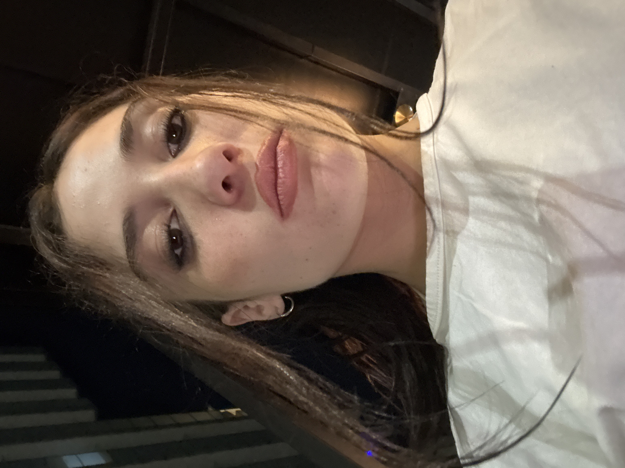A selfie shows a person with light skin, brown hair, and dark eyes, wearing a white t-shirt. They have a hoop earring in their left ear and are looking slightly downward. The background suggests an outdoor setting at night with urban elements, possibly in Buenos Aires, Argentina, indicated by the style of the architecture visible.