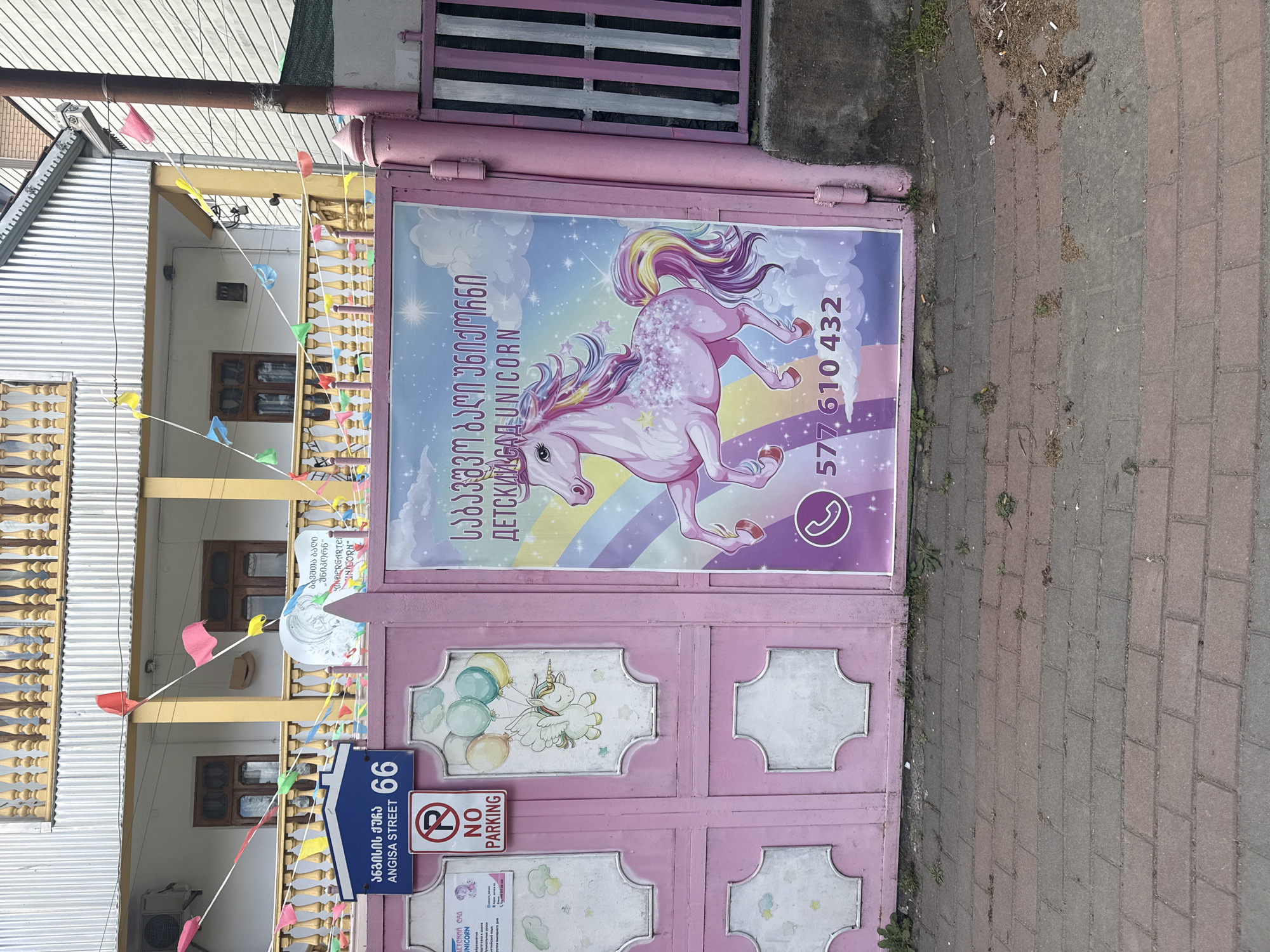The image shows the exterior of a building in Batumi, Georgia. The building appears to be a kindergarten or childcare facility based on signage and decorations. The building has a second-story balcony adorned with colorful pennant flags. A pink metal gate in front of the building displays signage with a unicorn design and the words "UNICORN", "NO PARKING", the Georgian text "საბავშვო ბაღი უნიქორნი/ДЕТСКИЙ САД UNICORN", the phone number "577 610 432", and the street address "ANGISA STREET 66". The sidewalk is paved with light grey bricks.